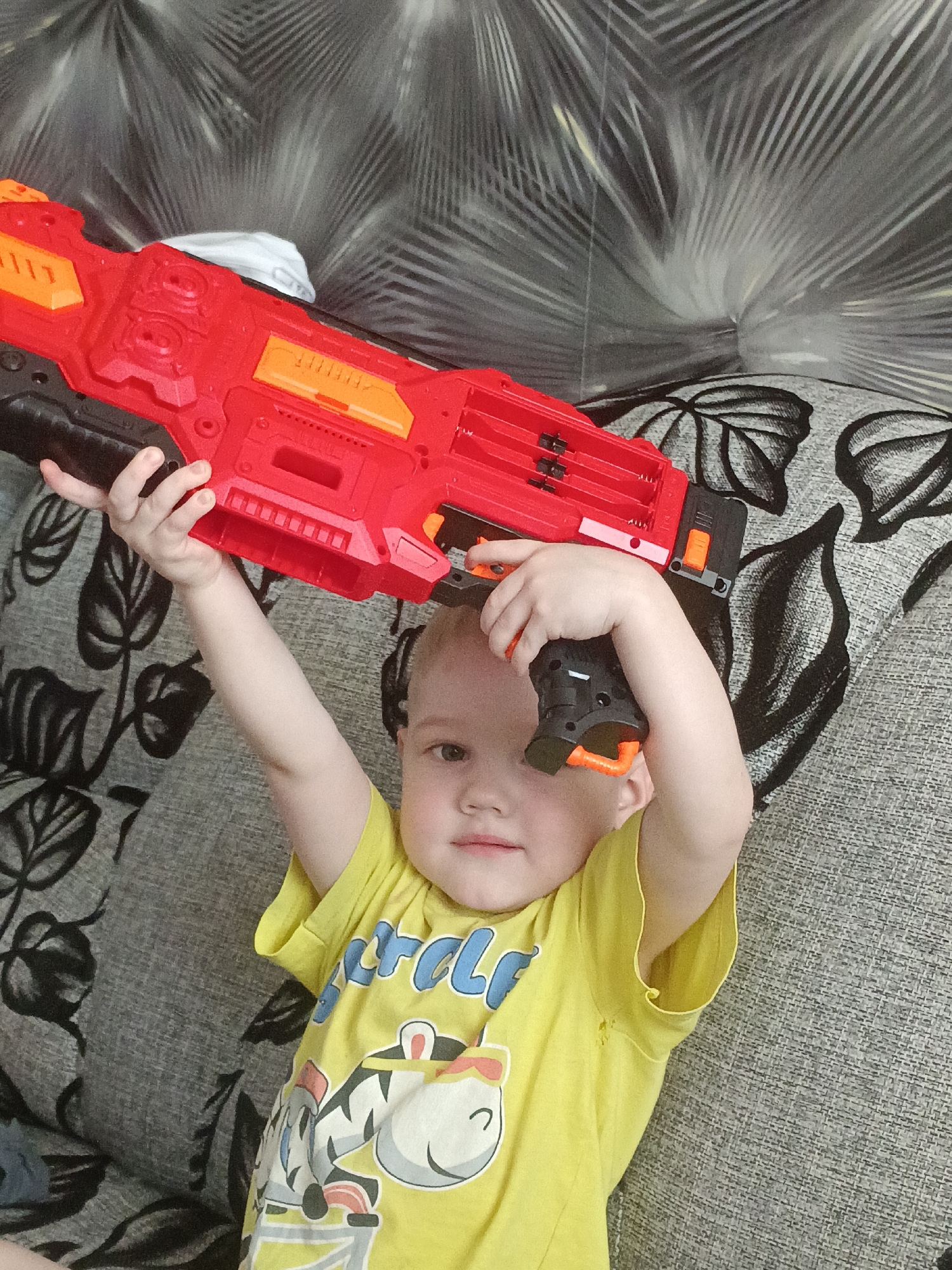 A young child, appearing to be under the age of five, is lying down on a patterned sofa. The child is wearing a yellow t-shirt with a cartoon image on it. The child is holding a large red toy gun above their head with their right arm. The background consists of the sofa and a wall with a textured pattern. The image is a casual, indoor photograph. No specific location in Chaikovsky, Russia, can be determined from the visual information provided.