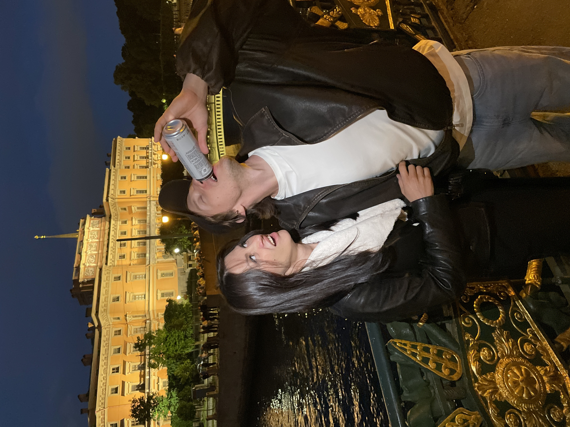 In Saint Petersburg, Russia, at night, a couple stands on a bridge. A man, wearing a leather jacket, white t-shirt, and blue jeans, drinks from a can. A woman, in a leather jacket and scarf, looks at him. Behind them, a body of water reflects the lights of a building. The building, a grand palace with illuminated windows and a spire, is in the background. The bridge is adorned with ornate golden railings. Trees and a nighttime sky complete the scene.