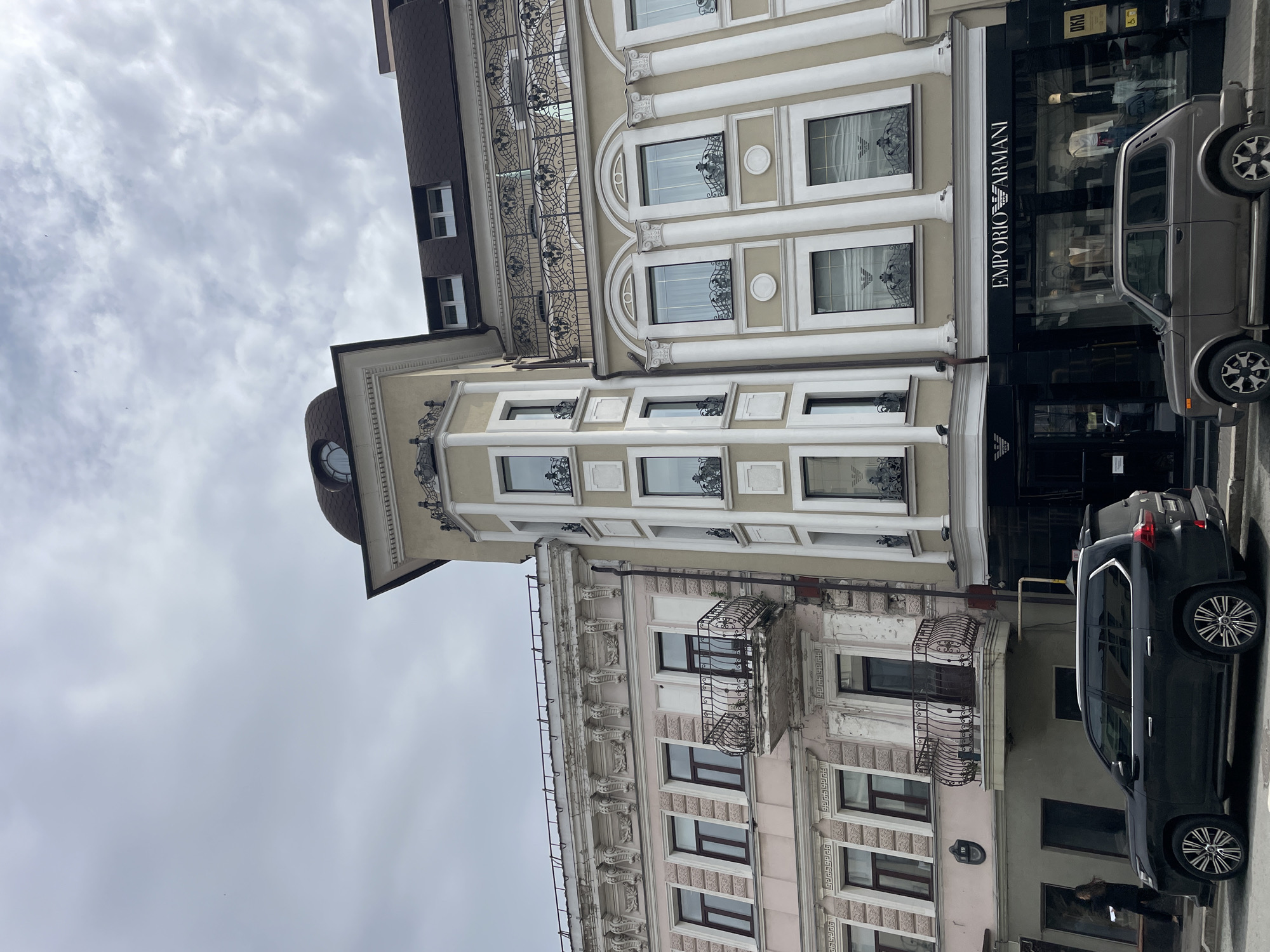 The image shows a street scene in Kazan, Russia. Two buildings are visible, one with an Emporio Armani store at street level. A gray Lada Niva and a dark SUV are parked in front of the buildings. A person is standing on the sidewalk. The sky is cloudy. The buildings have ornate architectural details.