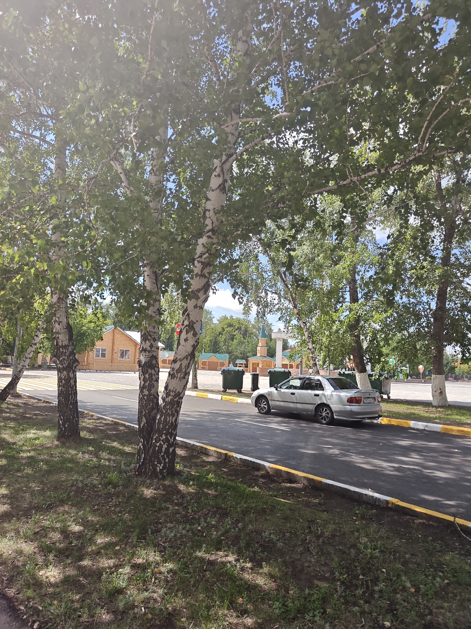 The image depicts a street scene in the city of Petropavl, Kazakhstan. In the foreground, several birch trees with distinctive white and black bark line a paved road. A silver sedan is parked on the right side of the road, facing away from the camera. The car's license plate is visible and reads "C817AA 15". Behind the trees on the left, a small wooden building with a brown roof and windows can be seen. Further in the background, there are more trees, some buildings with green roofs, and several large green garbage bins. The sky is visible in the upper part of the image, with patches of blue and white clouds. The overall impression is one of a sunny day in a park-like urban setting.