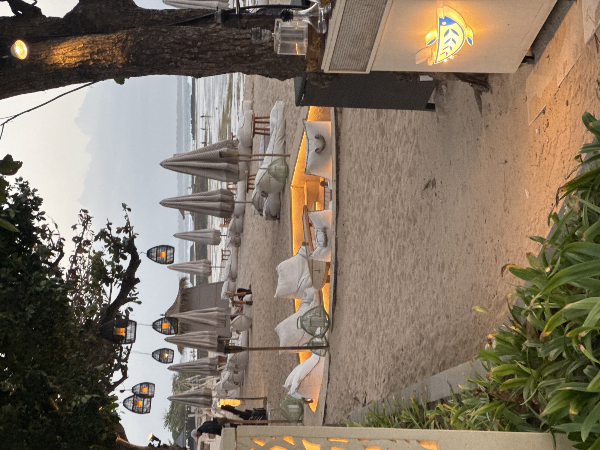 This image depicts a beachside seating area in Kuta Selatan, Indonesia, at dusk. In the foreground, a paved walkway borders lush green plants. The sandy beach is furnished with numerous white beanbag chairs arranged around a sunken, illuminated circular seating area. Several thatched umbrellas are set up on the beach further back, with more beanbag chairs and small wooden tables. Hanging lanterns with orange lights are suspended from tree branches overhead. In the middle ground, people are visible on the beach and wading in the shallow ocean water. The background shows the ocean meeting a hazy sky. To the right, a beige structure with a prominent, illuminated sign featuring a blue fish and olive branches is visible. Various bottles and what appears to be a hookah are placed on top of the structure. The overall ambiance suggests a relaxed, tropical evening setting at a beachfront establishment.