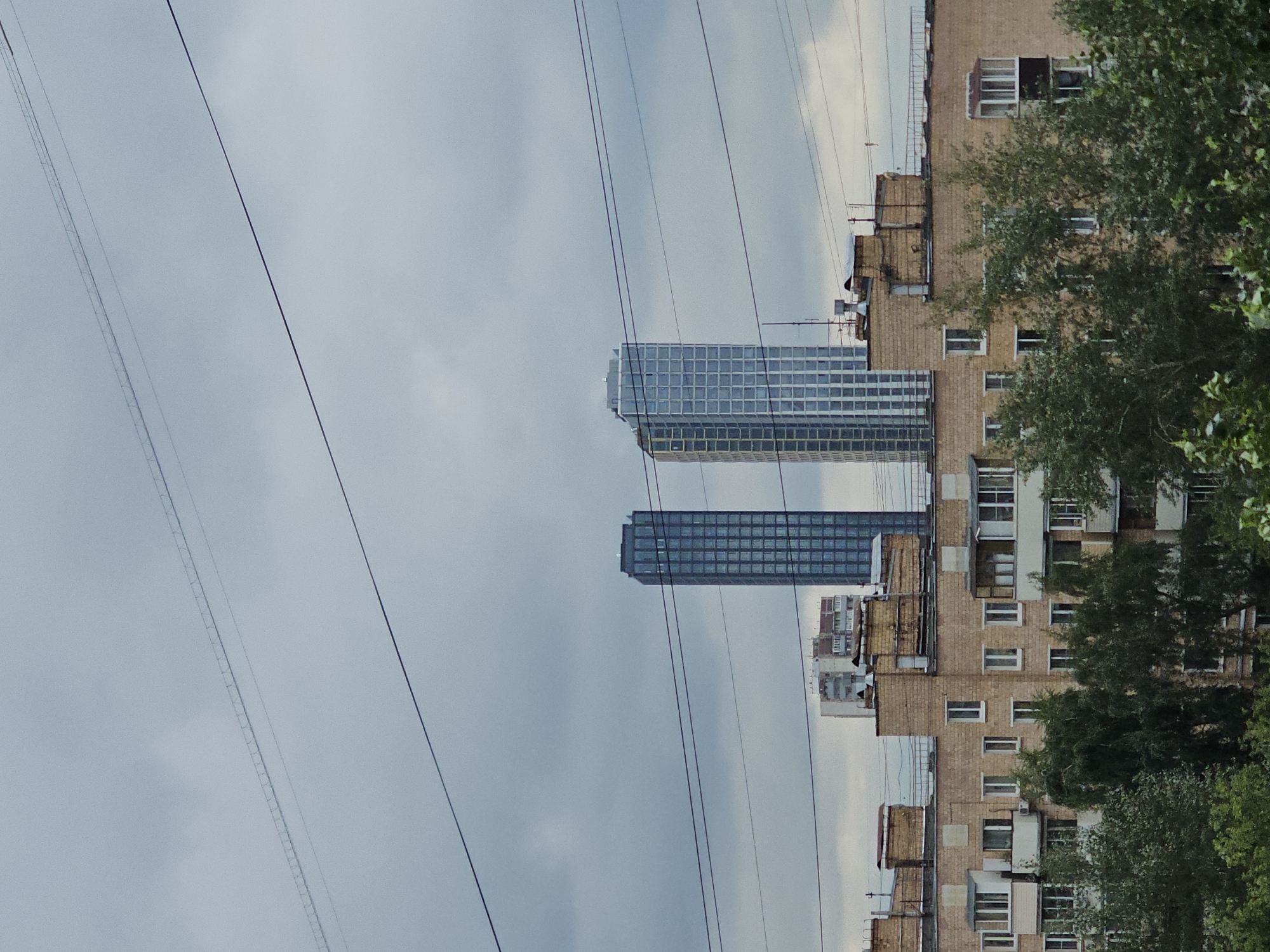 The image shows a view of apartment buildings and skyscrapers in Moscow, Russia. The foreground features a section of a mid-rise apartment building with a beige brick facade and multiple windows and balconies. Dense green trees are in front of and partially obscuring this building. In the background, two modern, glass-covered skyscrapers stand prominently against a cloudy sky. Another, lighter colored apartment building is visible between the foreground building and the skyscrapers. Several power lines are strung across the frame, running horizontally at different heights, indicating an urban environment with overhead electrical infrastructure. The sky is overcast, suggesting a cloudy or possibly rainy day.