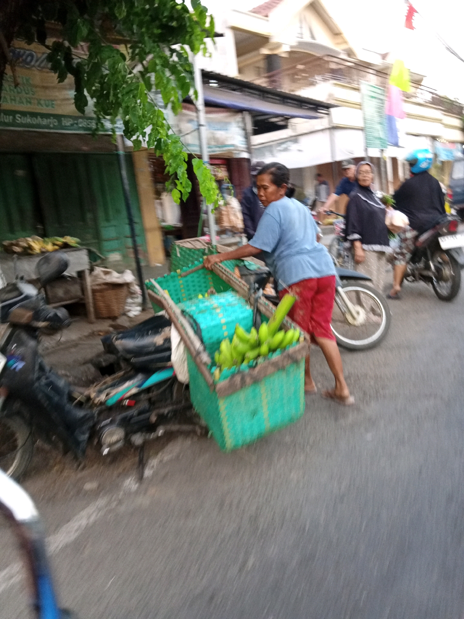 An outdoor, eye-level, medium shot captures a street scene in Bendosari, Indonesia, likely during the daytime. A woman wearing a blue t-shirt and red shorts is the central focus, pushing a cart filled with bright green bananas. The cart itself is a woven, teal basket, also carrying other unidentifiable items. Her body is slightly blurred due to motion, suggesting she is actively moving.
Behind the woman, a dark-colored motorcycle is parked, partially obscured by the banana cart. Further back, other individuals are visible, including a person on a motorcycle wearing a blue helmet and a dark jacket, and another woman in a black headscarf and shirt. The background reveals buildings with awnings and signs, consistent with a marketplace or commercial area. Lush green foliage hangs from a tree branch in the upper left corner, partially obscuring a sign with Indonesian text. The ground is asphalt, indicating a road or paved area. The overall impression is of a bustling, everyday moment in a local Indonesian community.