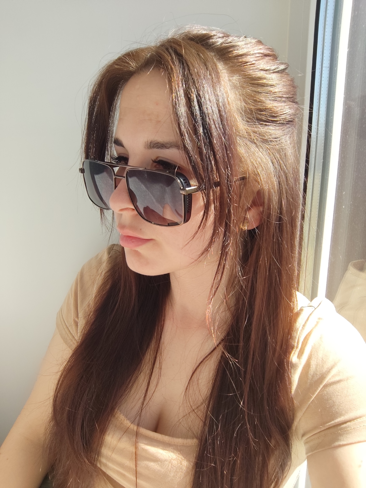 A woman is depicted in a medium shot. She has light brown hair with darker brown lowlights, and her hair is styled with some sections pulled back from her face. She is wearing large square-frame sunglasses with dark lenses. She is wearing a light beige or tan T-shirt. Her expression is thoughtful, and she is looking off to the left side of the frame. She has light skin and full lips.
The background is a neutral white wall and a window with sunlight streaming in. The location of Фоки, Russia is not discernible from the image.