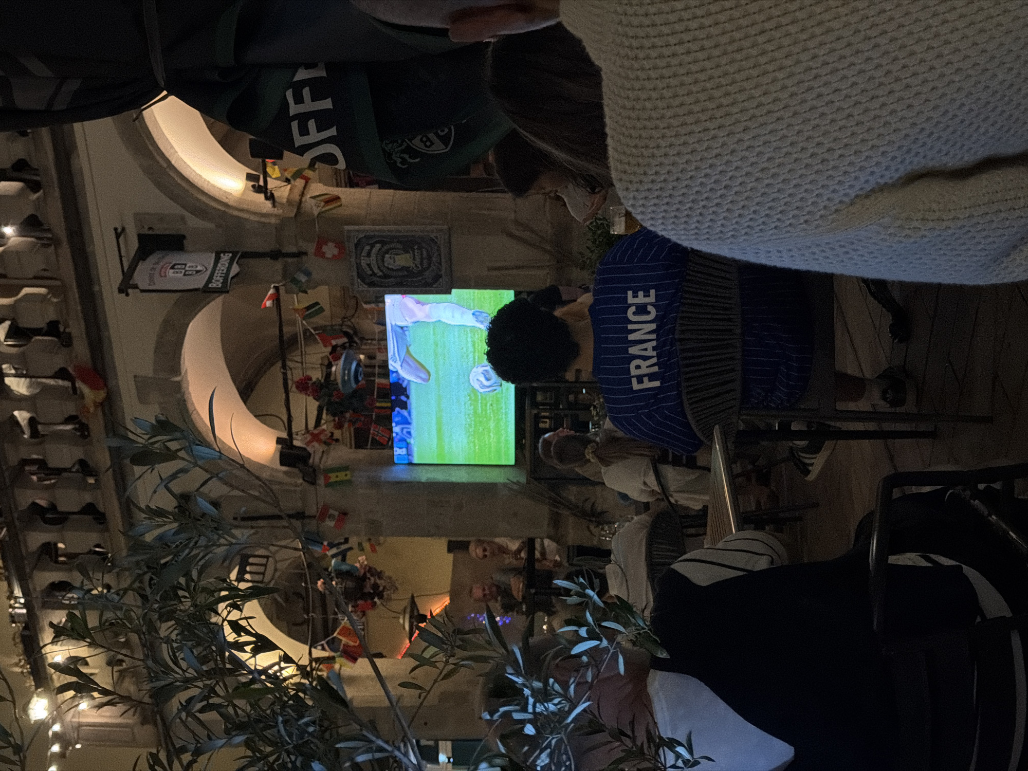 The image captures an outdoor seating area of a establishment, likely a bar or restaurant, in Luxembourg City. A large screen displays a soccer match, with the green turf and players' legs visible. Several people are seated at tables, some facing the screen, others in conversation. In the foreground, a person wearing a blue and white striped jersey with "FRANCE" printed on the back is seated. To the right, another person is partially visible, wearing a light-colored textured sweater. The background reveals arched architecture and string lights, suggesting an evening or nighttime setting. Small flags of various nations are strung across the area, adding to the festive atmosphere often associated with sports events. The overall scene depicts patrons enjoying a soccer match in a relaxed outdoor ambiance.