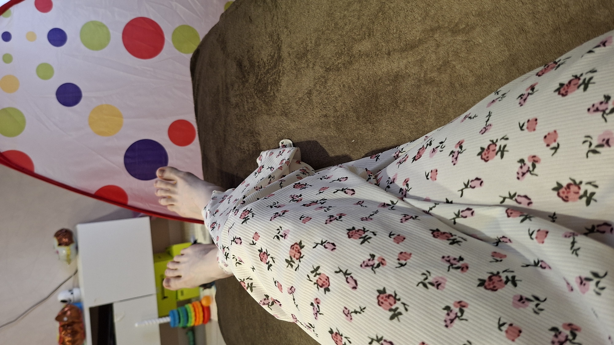 The image shows a person lying on a brown sofa. They are wearing white pajamas with a pink floral print. The person's bare feet are visible. In the background, there is a white and multicolored polka-dotted play tent, along with a white dresser and some toys.
