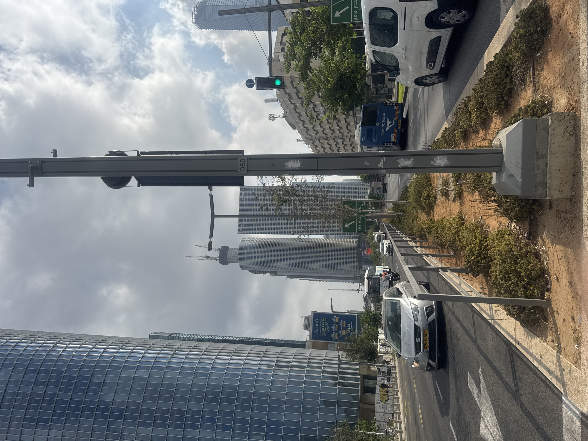 The image depicts a wide, multi-lane road in Tel-Aviv, Israel, with a prominent street light pole in the foreground. On the left, a tall, modern glass skyscraper dominates the scene, reflecting the cloudy sky. Several other skyscrapers, some under construction, are visible in the background. The road is busy with traffic, including cars, a bus, and a motorcycle. A silver car with the license plate "625 90 501" is in the left lane. A green traffic light is illuminated. The street is lined with metal railings and low green bushes. The environment appears to be an urban business district with contemporary architecture.