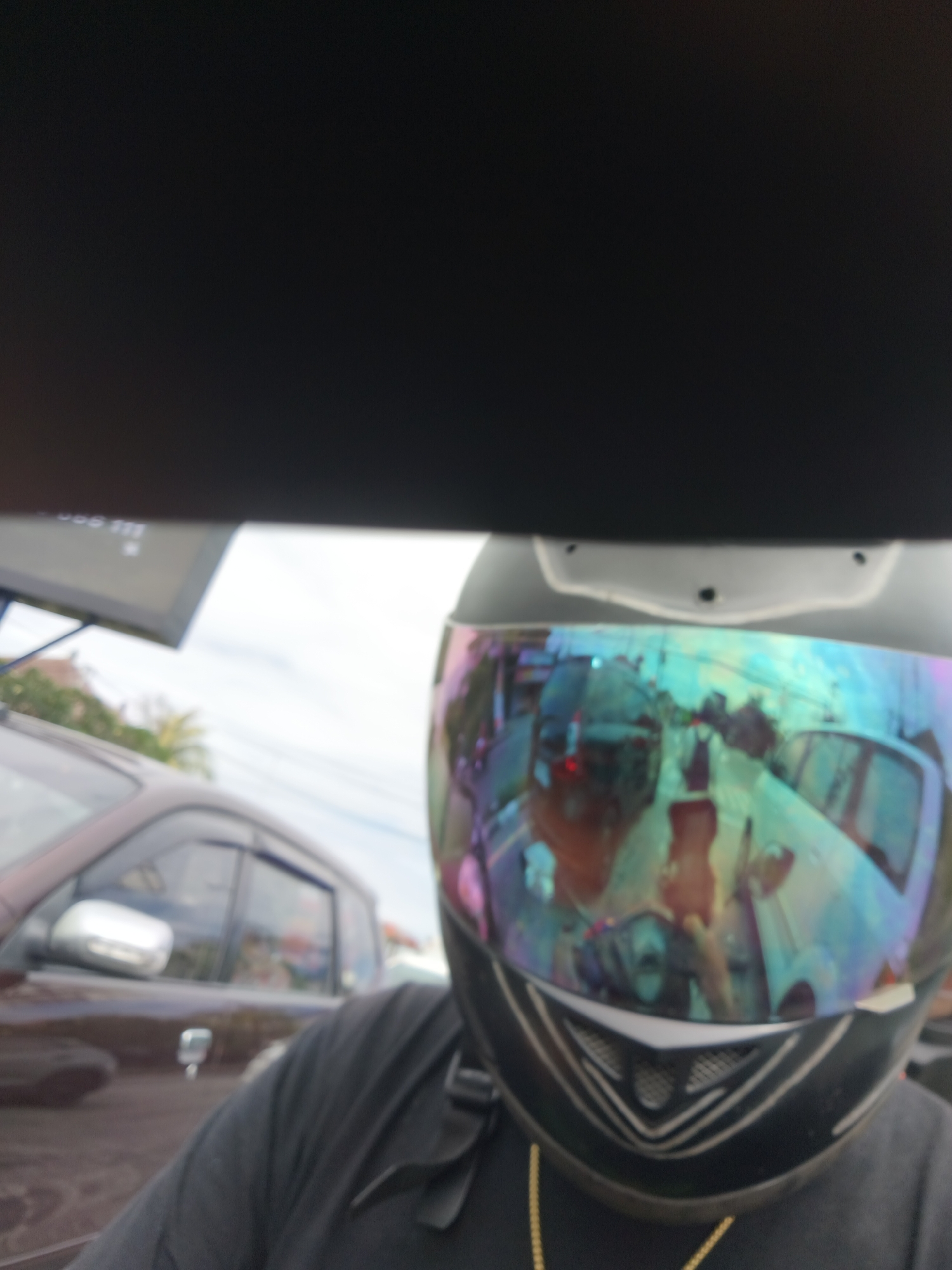 The image shows a person wearing a motorcycle helmet with a reflective visor. The visor reflects a street scene, appearing to be a busy road with several vehicles, including cars and motorcycles. The context of Denpasar, Indonesia, suggests a tropical or sub-tropical environment, possibly with dense traffic characteristic of urban areas in Southeast Asia. The time of day appears to be daytime, based on the lighting conditions. The person's clothing, a dark t-shirt, is partially visible. The reflection in the visor shows the person's face, distorted by the helmet's curvature, and the surrounding environment, including buildings and vegetation. The scene captured in the reflection is a typical street view, with other vehicles on the road and possibly pedestrians. The overall image is a selfie taken from the perspective of the motorcyclist.