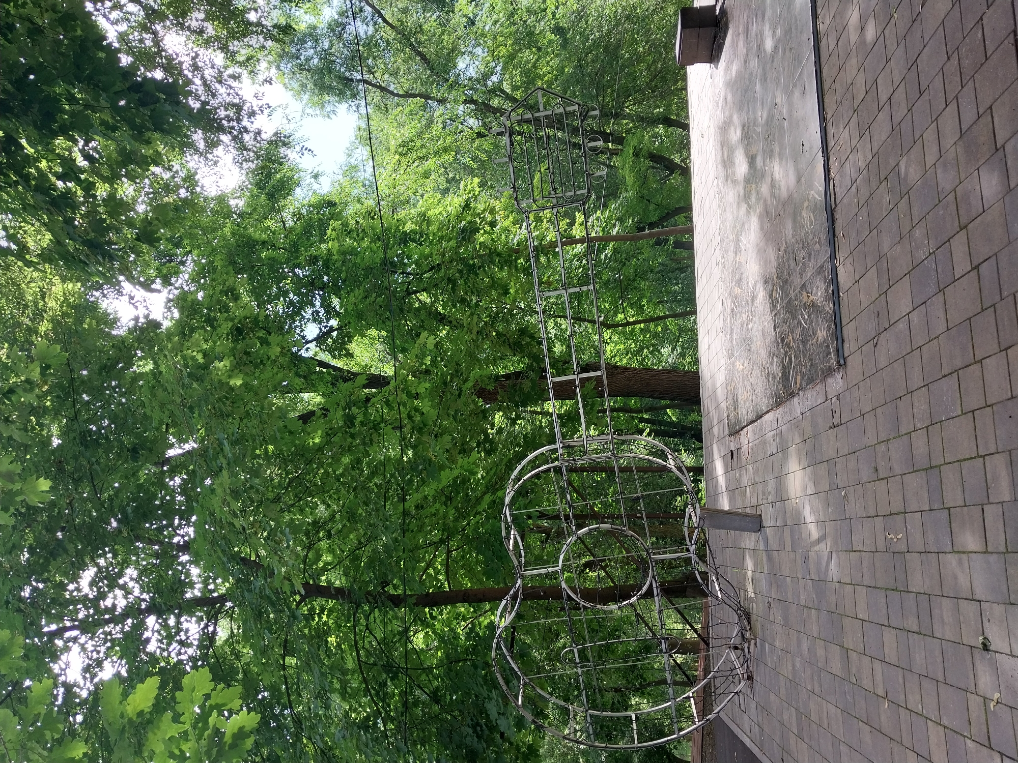 In Dnipro, Ukraine, an outdoor scene depicts a large, skeletal sculpture resembling a guitar made of metal framework, positioned on a paved area. The sculpture is situated in front of a dense forest with lush green trees, suggesting a park or natural area. The foreground features a square, grey, brick-patterned pavement, transitioning to a section of larger, flatter grey tiles. Sunlight filters through the trees, creating dappled shadows on the ground and sculpture. The overall atmosphere is serene and natural, with the metallic structure providing a modern contrast to the organic surroundings. There are no people or animals visible in the image.