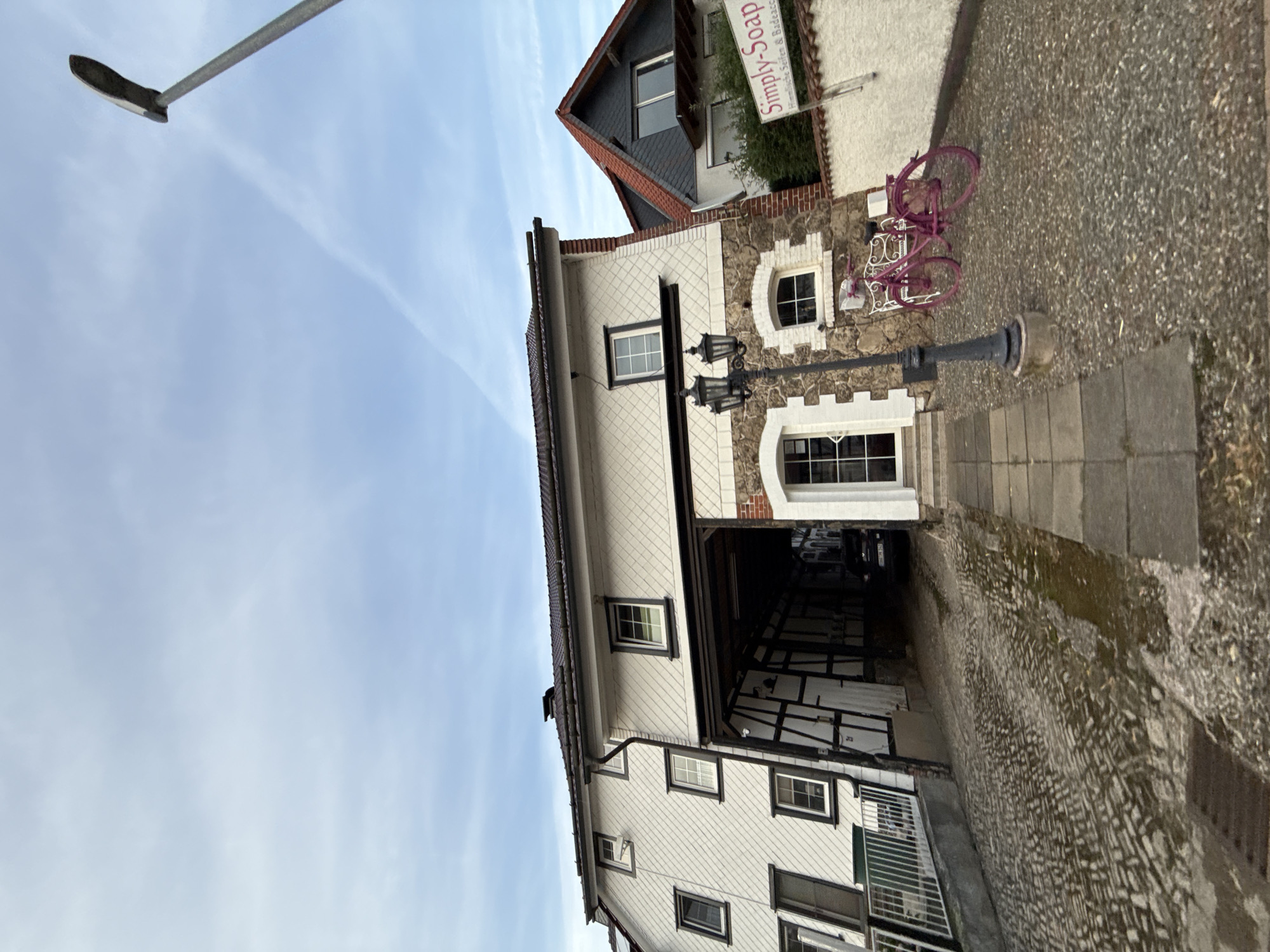 The image depicts a quaint street scene in the city of Bad Sachsa, Germany. The foreground features a cobblestone street that appears wet, possibly from recent rain, with a narrow paved walkway to the right. To the left, a white building with diamond-shaped siding and dark-trimmed windows is visible. An arched entryway, supported by dark wooden beams, leads into a covered area where a dark car is parked. Adjacent to the entryway, a decorative black lamppost stands in front of a building with stone and white-trimmed windows. A bright pink bicycle is parked on a gravel area in front of a white wall on the right side of the image, and a sign reading "Simply-Soap" is mounted on the wall above some greenery. The sky above is cloudy with streaks of lighter clouds. The scene conveys a sense of a quiet, European town.