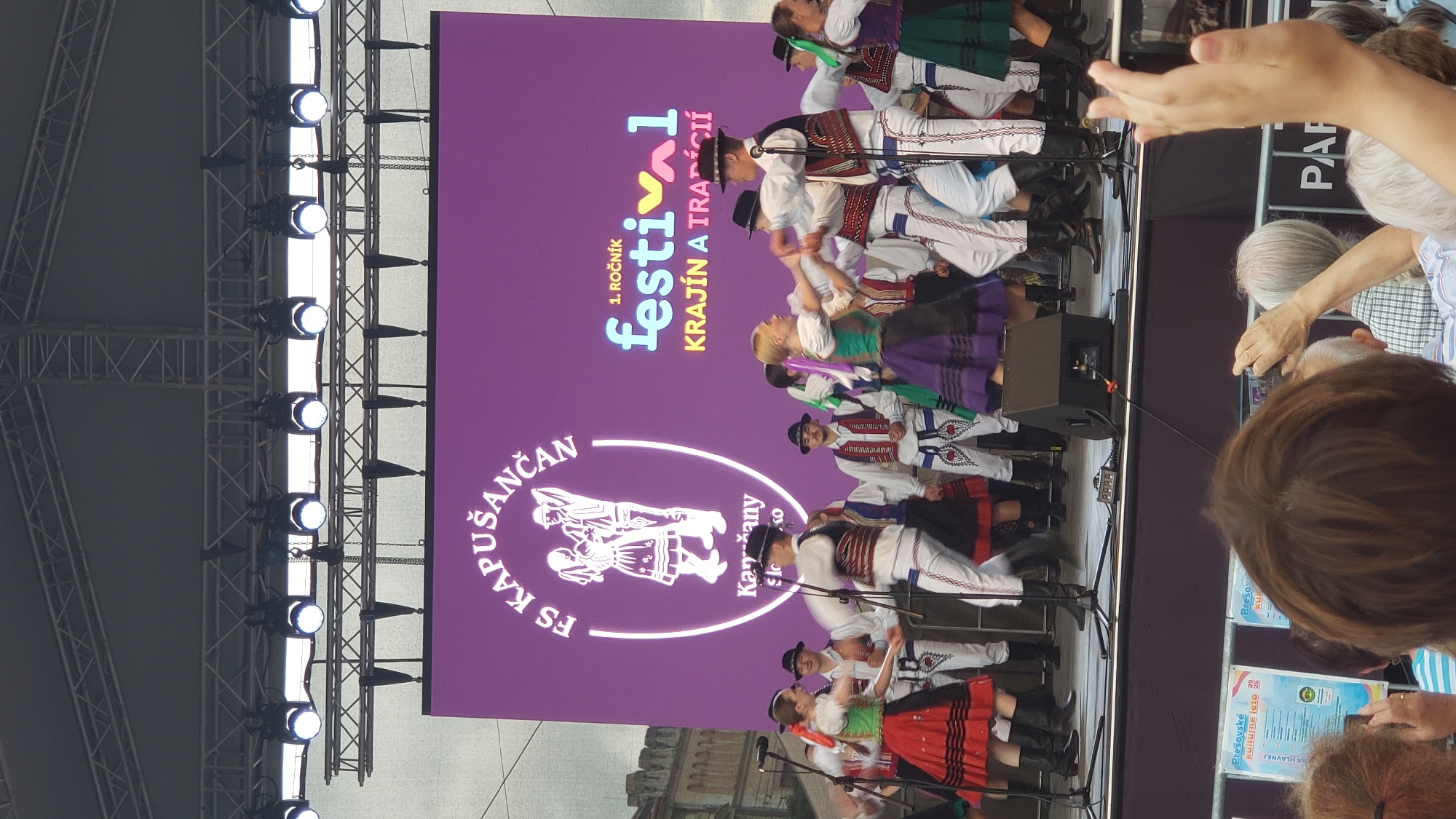The image depicts an outdoor performance by a folk dance ensemble on a stage in Prešov, Slovakia. A large purple LED screen is visible behind the performers, displaying the text "FS KAPUŠANČAN" and "FESTIVAL KRAJÍN A TRADÍCII" along with graphical elements. The ensemble consists of dancers in traditional Slovak attire, including women in red and green skirts and embroidered vests, and men in white pants, vests, and hats. Some dancers are actively engaged in a dance, with couples holding hands and moving. The stage is equipped with microphones and speakers. In the foreground, members of the audience are visible, some holding up phones to take pictures or videos. The overall atmosphere appears to be festive and cultural, likely part of a local festival or event.
