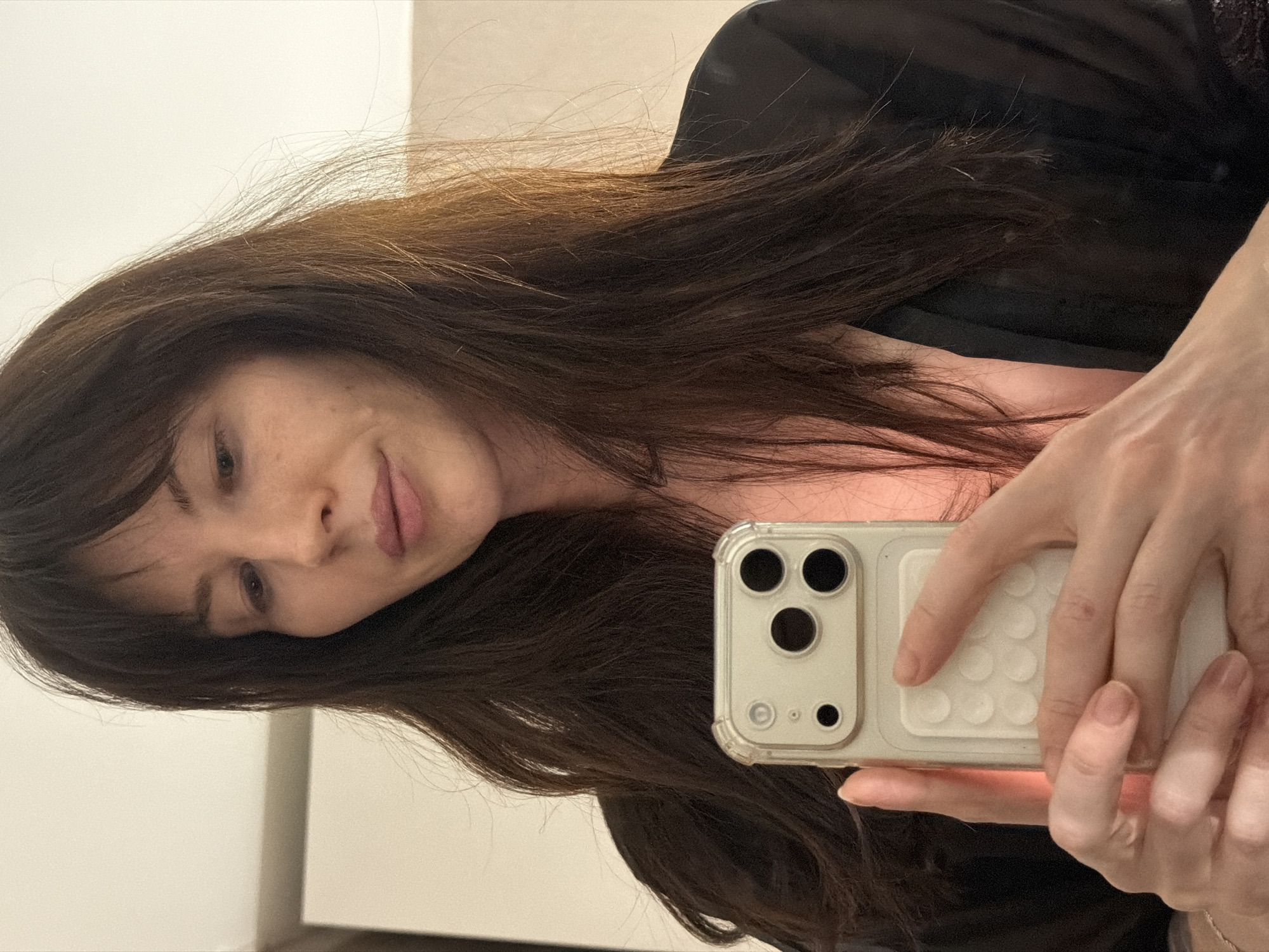 A woman is holding a cell phone and taking a selfie indoors. She has brown hair and is wearing a dark-colored garment. The environment appears to be a room with a white wall in the background. The location information (Perm, Russia) cannot be verified from the image.