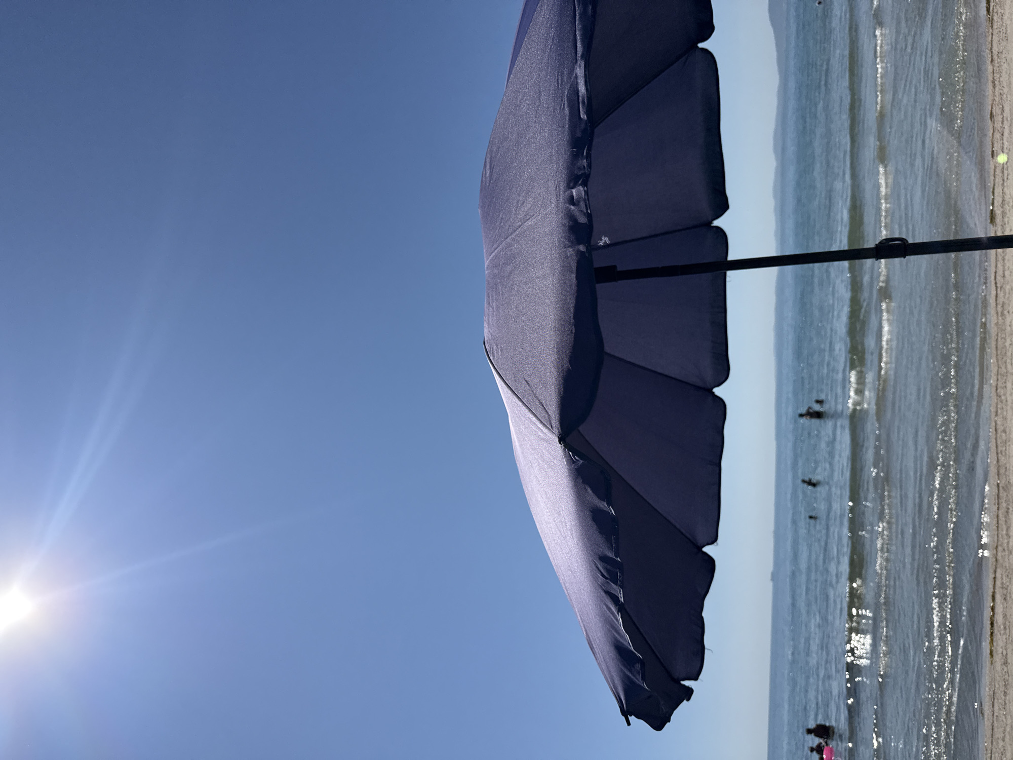 The image depicts a sun-drenched beach scene with a dark blue beach umbrella in the foreground, partially obscuring the view. The umbrella's fabric is textured and catches the sunlight. Beyond the umbrella, the calm, sparkling sea stretches towards a hazy horizon line where distant hills or mountains are visible. The sky is a clear, bright blue, with the sun intensely shining from the upper left corner, casting lens flares and rays of light. Several people are visible in the water, appearing as small silhouettes enjoying the sea. Some are wading, while others are further out. The water has gentle waves lapping at the sandy shore in the foreground. The overall impression is one of a peaceful summer day at the beach, likely in Pomorie, Bulgaria, known for its coastal location and sunny climate.