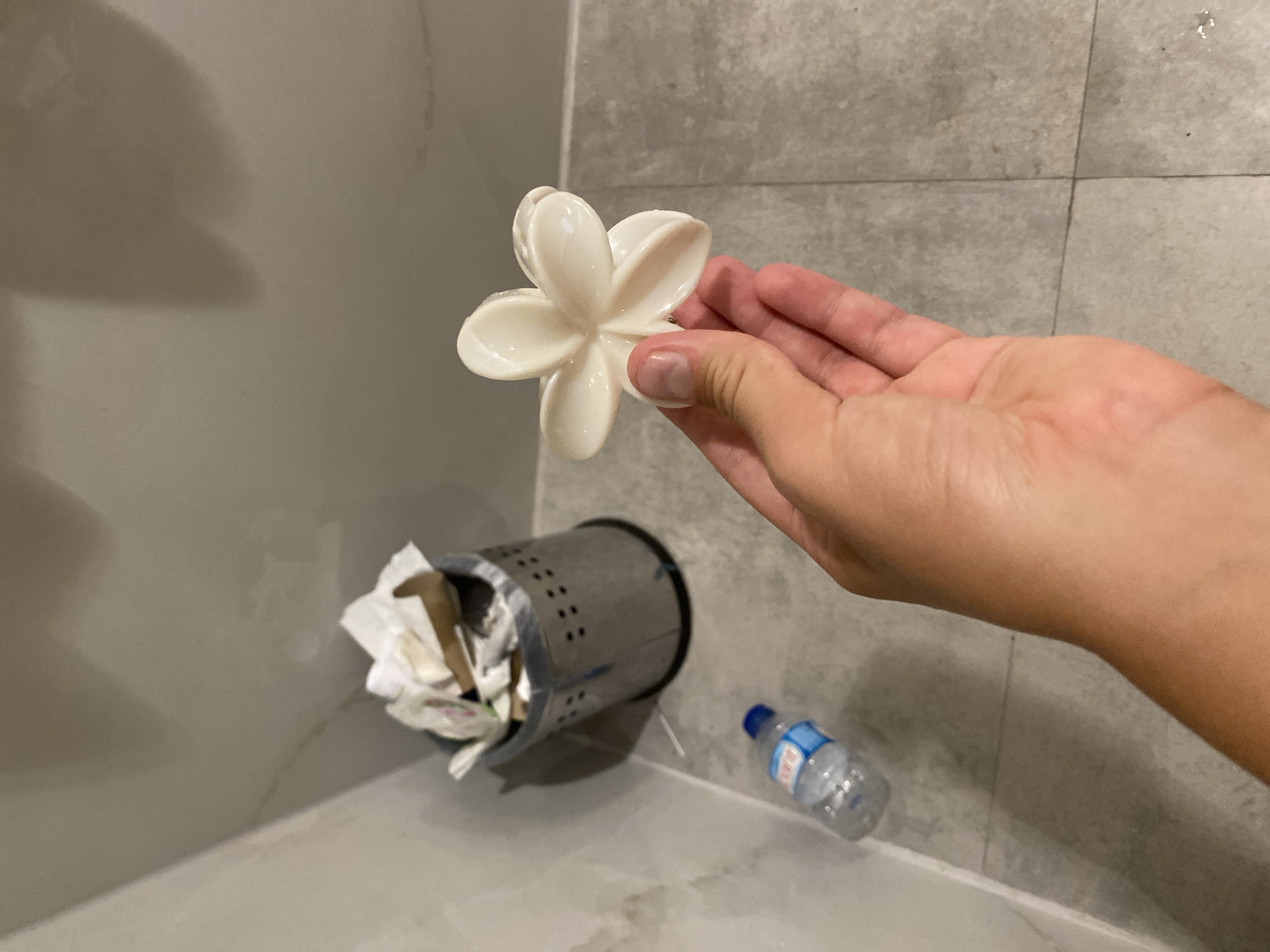 A first-person perspective image shows a hand holding a white, flower-shaped object. In the background, a silver trash can is filled with crumpled paper. To the left of the trash can, a small plastic bottle of water lies on the floor. The environment appears to be a bathroom or restroom, with tiled walls and floor. The image does not provide any information about the city of Aksu, Turkey.