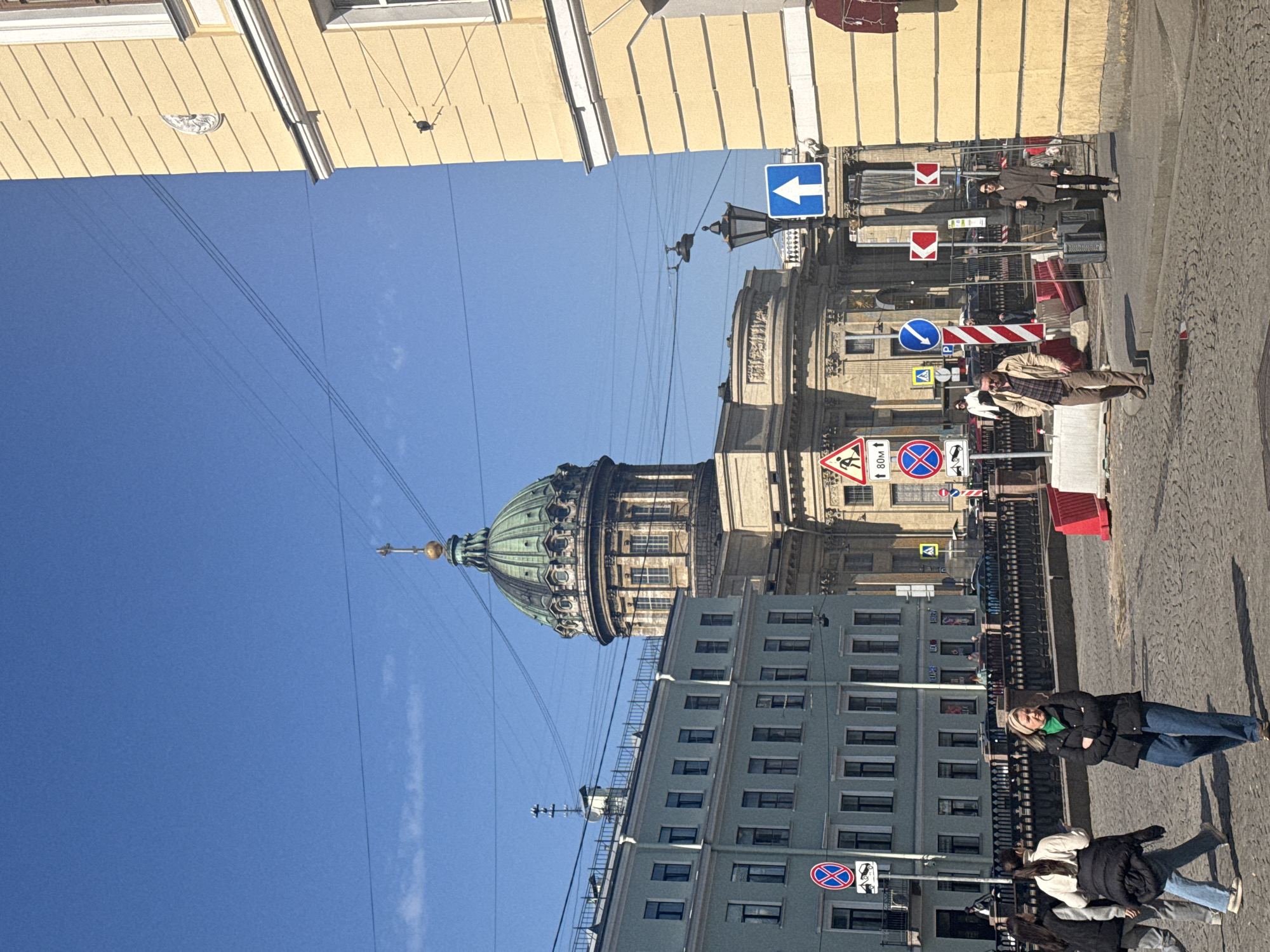This image captures a vibrant street scene in Saint Petersburg, Russia, under a clear blue sky. Dominating the background is the iconic, ornate green dome of the Kazan Cathedral, its intricate architectural details clearly visible against the bright sky.
The foreground features a bustling city street lined with buildings and several pedestrians. On the left, a light yellow building with visible window frames runs along the top edge of the frame. On the right, a multi-story building with a light blue-grey facade stands, featuring numerous rectangular windows.
Several individuals are visible on the cobblestone pavement. A woman with blonde hair, dressed in a black jacket, green top, and blue jeans, stands near a metal fence. Beside her, another person in a white jacket and jeans is partially visible. Further down the street, a man wearing a light brown jacket and a plaid shirt is present, and another woman in a brown coat and dark pants stands near red and white striped construction barriers.
The street is equipped with various traffic signs: a blue directional arrow pointing left, a red "No Entry" sign, a blue circular sign indicating "ahead only" with a white arrow pointing right, a red triangular "road works" sign, and multiple "No Stopping" signs. One "No Stopping" sign includes Russian text that translates to "Vehicles of persons with special passes 🦽" (for disabled access). Another white rectangular sign displays "80M ⬆️". Overhead power lines crisscross the sky.
The time of day appears to be daytime, characterized by bright sunlight casting visible shadows. The weather is clear and sunny, contributing to a lively urban atmosphere.