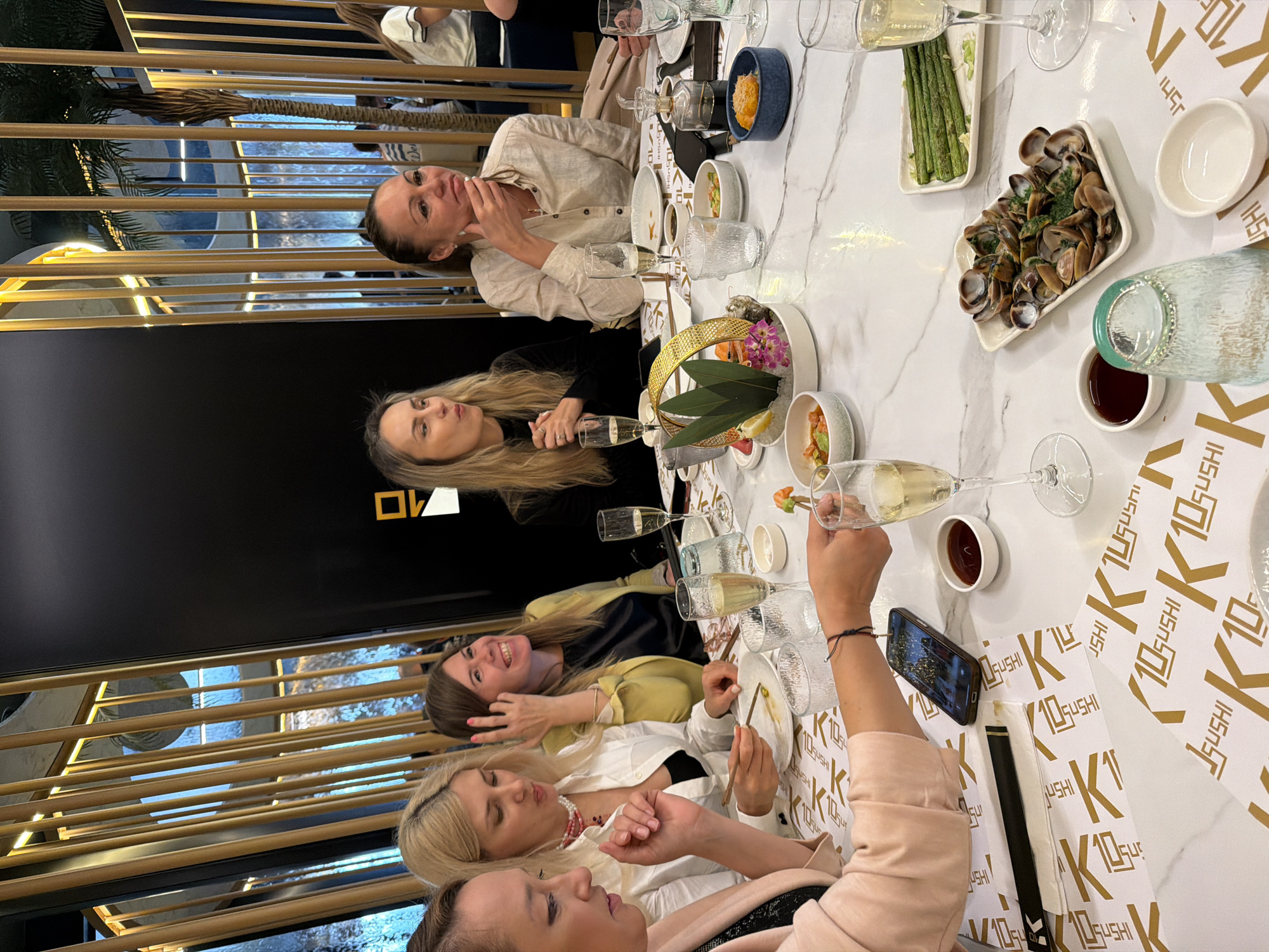 Here is a concise description of the image:
**Scene:** The image captures a group of women enjoying a meal at a restaurant. The table is set with several dishes, including what appears to be sushi, asparagus, and clams, along with accompanying condiments and glasses of champagne. The table is covered in white paper with a logo or design that may be related to the restaurant.
**People:** Several women are seated at the table, engaging in conversation. One woman is holding up her smartphone, possibly taking a photo or video of the scene.
**Environment:** The interior of the restaurant includes a gold decorative element with horizontal lines.