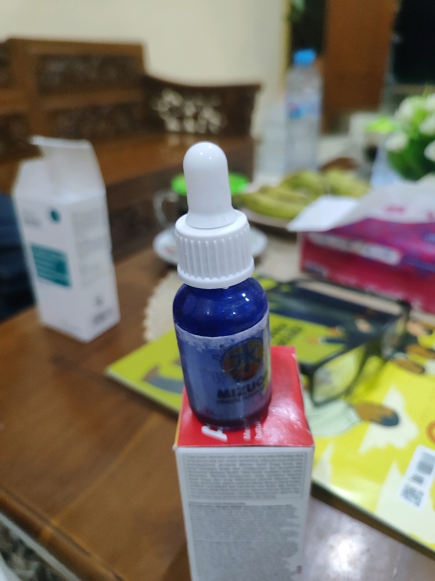 A blue dropper bottle with a white cap sits on top of a red and white box on a wooden table. The bottle is partially obscured by a pink and white object. In the background, a white container and a blurred bottle of water are visible. The scene appears to be indoors, possibly a home or office.