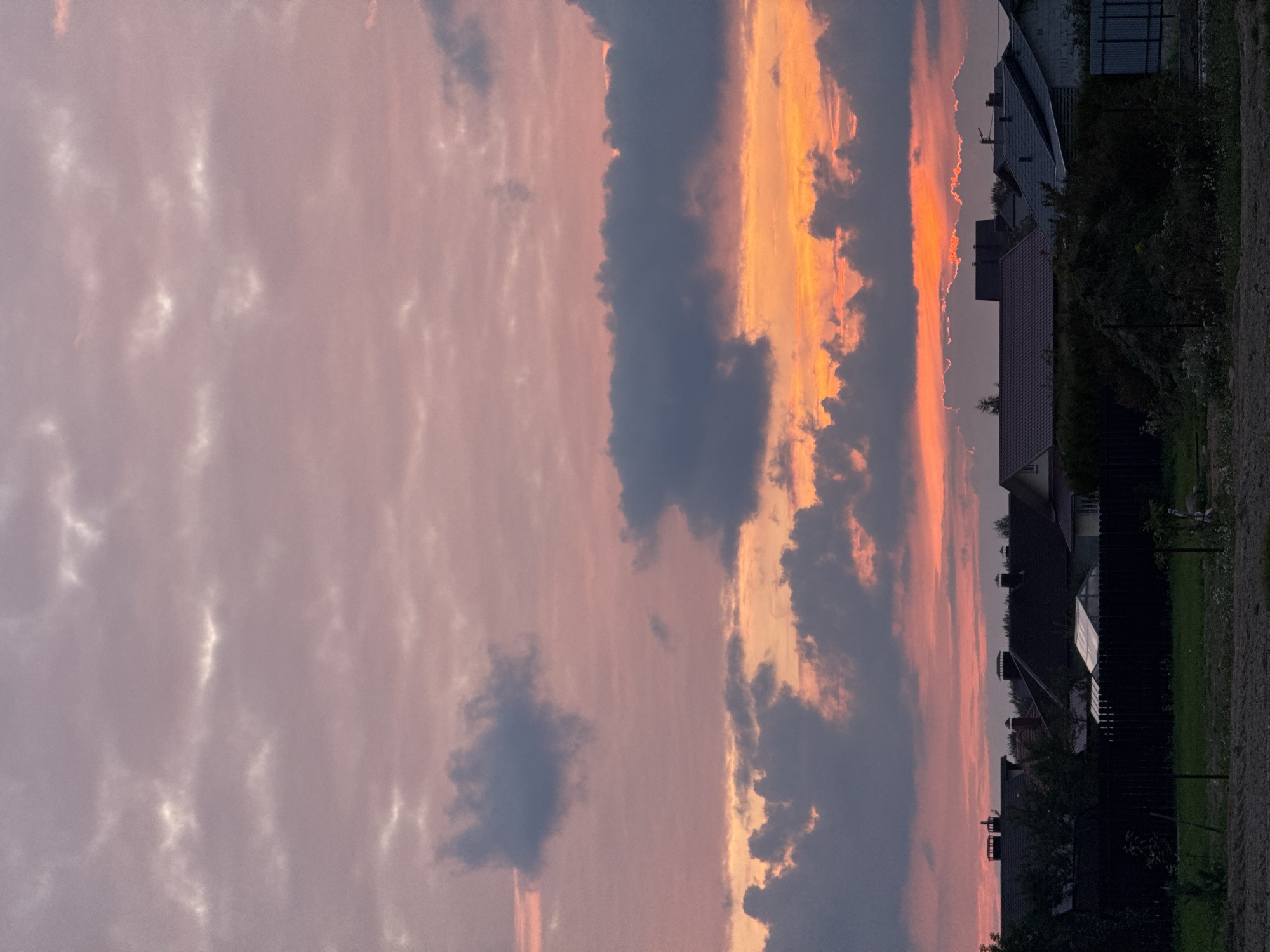 The image depicts a suburban or rural landscape at sunset in the Бараўлянскі сельскі Савет, Belarus. The sky is filled with dramatic clouds, transitioning from soft pinks and purples in the upper portions to vibrant oranges and yellows closer to the horizon, indicating the setting sun. Darker, gray clouds are interspersed throughout, with some appearing to be illuminated from behind by the sunset. Below the sky, a silhouette of houses with peaked roofs and chimneys is visible. A dark fence runs across the middle ground, with trees and vegetation behind it. In the foreground, there is a patch of bare earth or soil, with some scattered plants and grass. The overall atmosphere is tranquil and serene, characteristic of a rural evening. There are no people visible in the image.