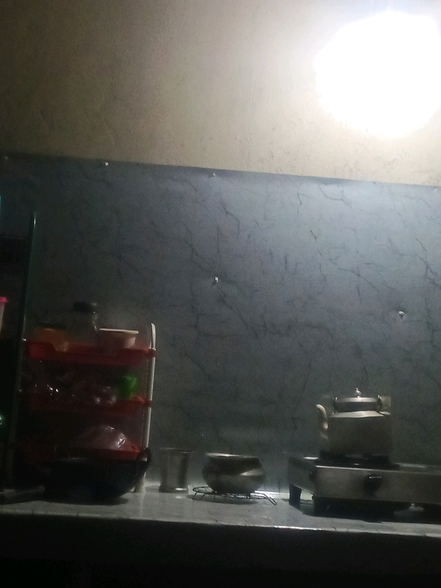 The image shows a dimly lit kitchen counter in Fulertal, India. On the left side of the counter is a red plastic shelf unit holding several bowls and containers. A dark pot is situated in front of the shelf. To the right of the pot, there are two metallic cups. Further to the right, a metallic kettle sits on a stovetop. The background consists of a grey, marble-patterned wall, with a bright light source illuminating the upper right corner of the image. The overall scene suggests a domestic setting, likely a kitchen.