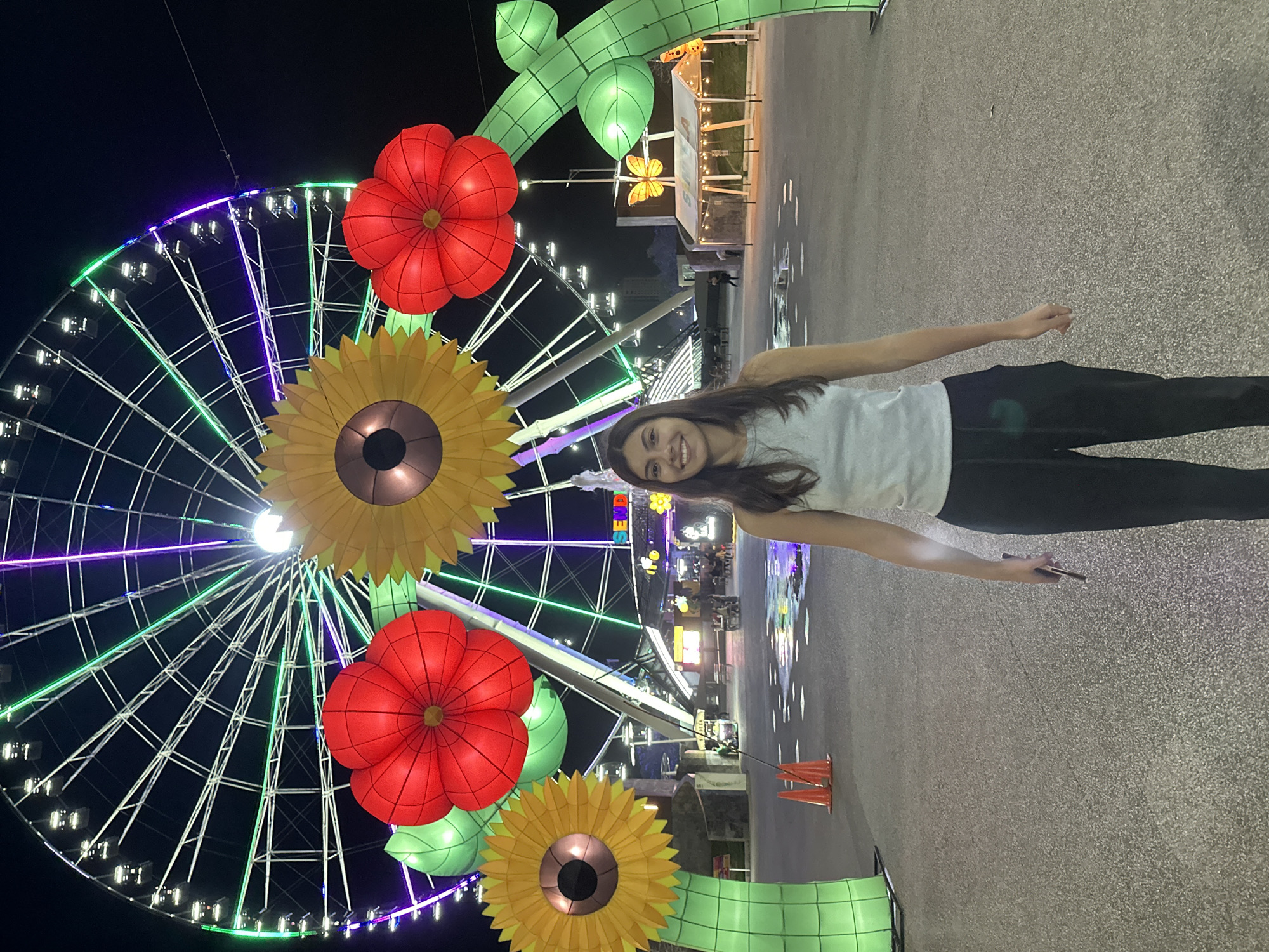 A medium-shot shows a young woman standing in front of a Ferris wheel at night, with artificial flowers decorating the foreground. The woman has long, dark hair and is wearing a white tank top and black pants. She is smiling and holding her arms out to the sides.
Behind her is a large Ferris wheel with green and purple lights. In front of the Ferris wheel are two large artificial flowers, a sunflower and a red poppy. The flowers are part of a larger decorative display with green stems and leaves. The ground is paved with a light-colored concrete. Further in the background, there are buildings and other attractions, also illuminated. The setting appears to be a festival or fair in the city of Puebla, Mexico.