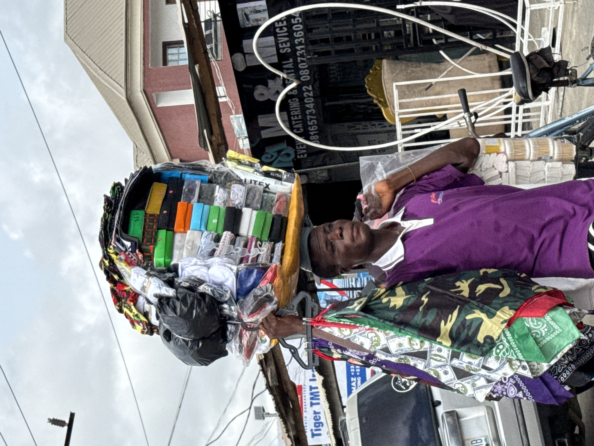 A man is carrying a large load of goods on his head and laying down, apparently to rest, on a bicycle piled with what appears to be rolls of fabric. The goods on his head include a variety of packaged items in different colors, possibly clothing or textiles. Additional fabric items, including bandanas with money prints and camouflage patterns, are stacked on the bike near him.
The scene appears to be outdoors, with buildings visible in the background. A sign reads "Tiger TMT LTD..." and "MAZ +234 / KING +234", suggesting a commercial or industrial area. Also in the background, a large sign says, "EVEN CATERING & FENTAL SEVICE / 08165734022 08073136054". The location can be further deduced from the text to be in Ado, Nigeria.
Overhead power lines are visible in the sky. The lighting is diffuse, suggesting an overcast day.