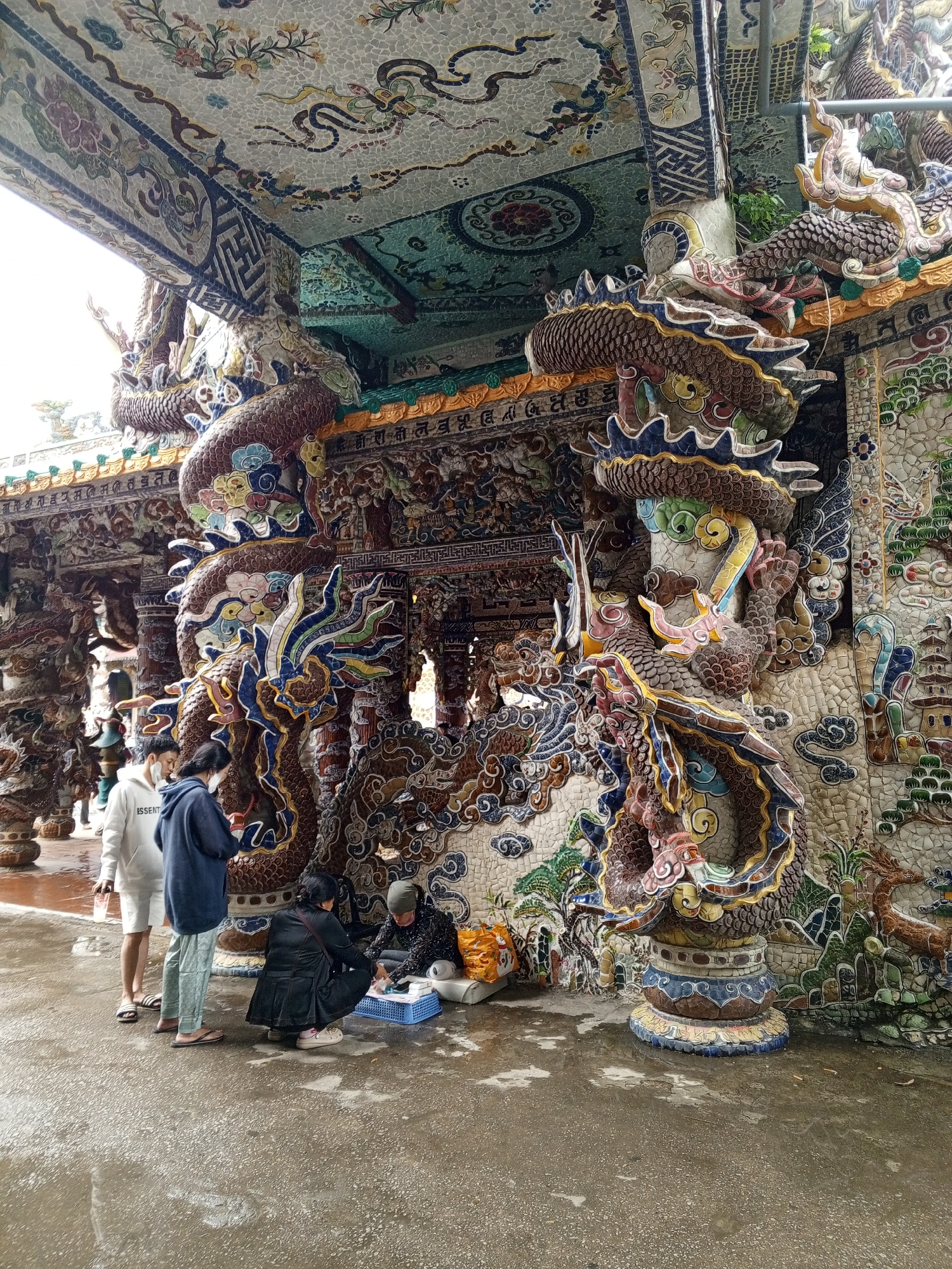 The image captures a scene in front of a structure adorned with intricate dragon sculptures and mosaic tilework, likely a temple or similar cultural site, possibly located in Xuân Thành, Vietnam. A group of people are gathered on a wet, paved ground surface. Three individuals stand to the left, two facing inward, engaged in conversation. One person, wearing a white hooded sweatshirt and shorts, holds a clear plastic cup. Another, in a dark blue hoodie and light green pants, has their hands in their pockets. A third person, partially visible, stands behind them. To the right, two individuals are crouched near a blue plastic basket. One, wearing a dark jacket, is bent over, possibly interacting with items in the basket. The other, in a patterned dark top and headscarf, is also crouched, appearing to be engaged in an activity with their hands. The background is dominated by the ornate architecture, featuring detailed mosaics of dragons, floral patterns, and mythical creatures. The ground is wet, reflecting the overcast sky, suggesting recent rain.
