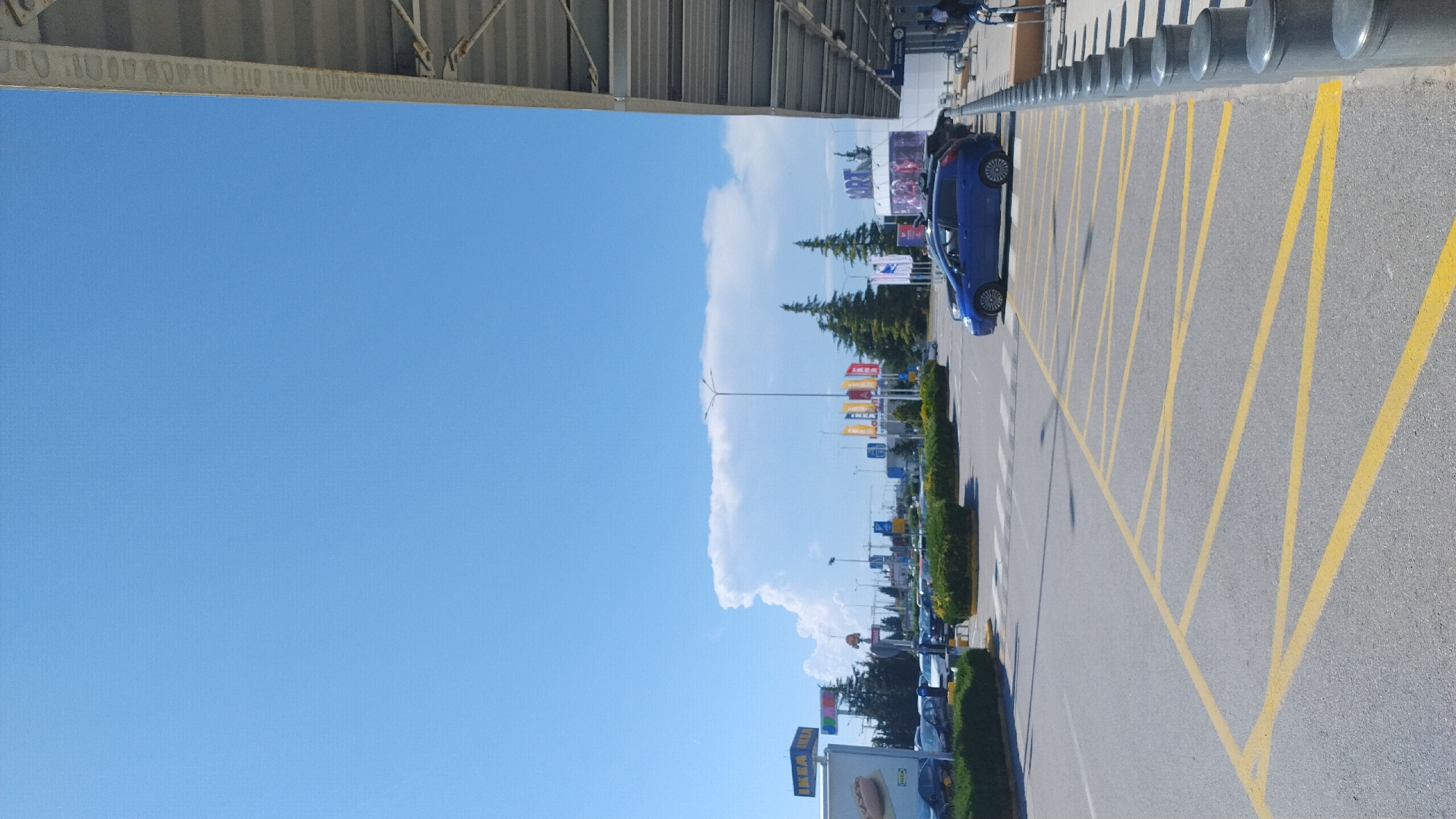 The image shows a bright, sunny day with a clear blue sky. In the foreground, a parking lot is visible with yellow lines painted on the asphalt. A blue car is parked on the right side of the lot, next to a row of bollards. Beyond the parking lot, there are several buildings with signage, including an IKEA store. There are also trees and some people walking in the distance. The scene appears to be a commercial area. The location is Pylaia Municipal Unit, Greece.