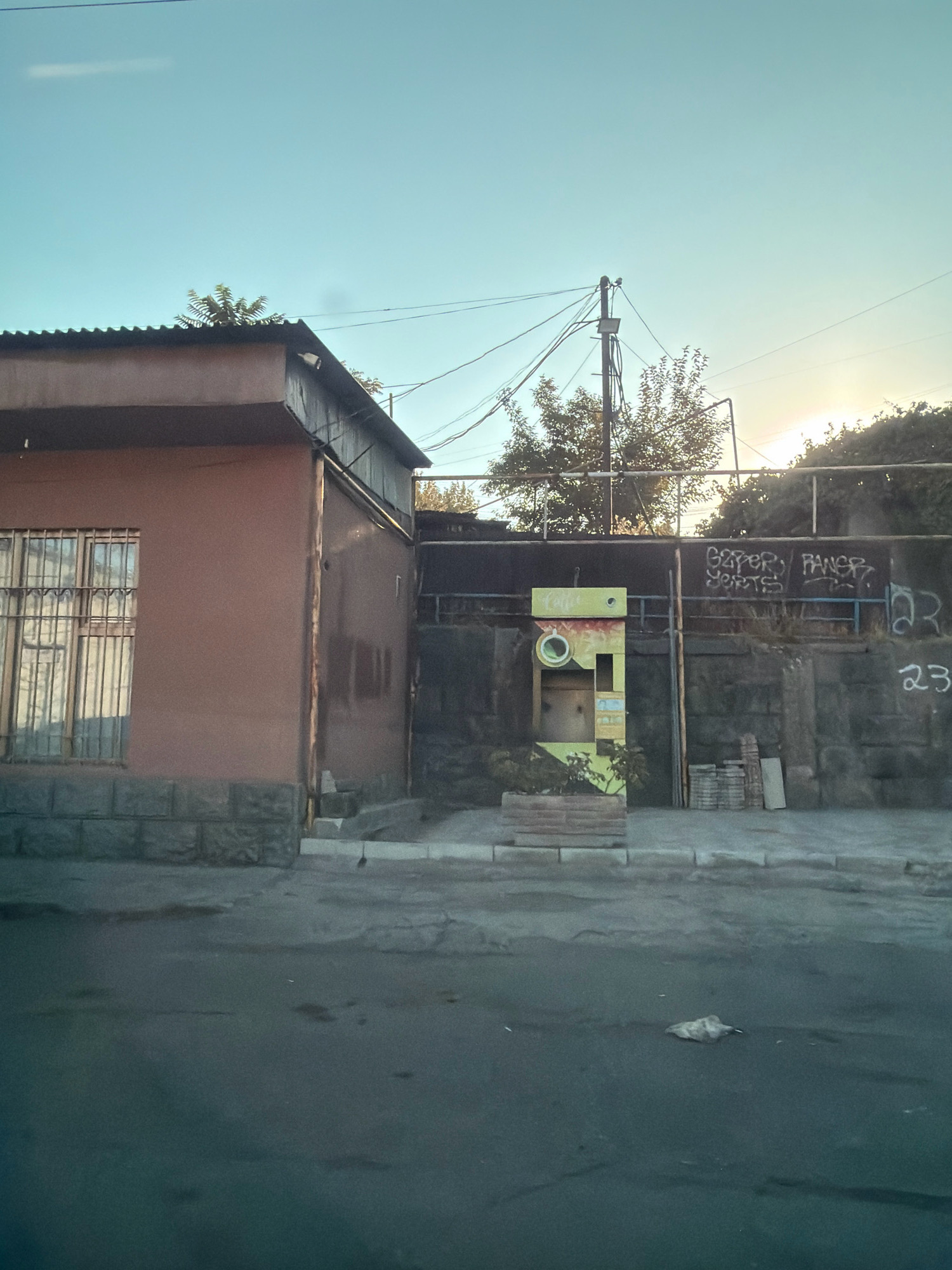 The image depicts a street scene in Yerevan, Armenia, under a clear, pale blue sky, likely during late afternoon or early morning given the low angle of the sun. In the foreground, a gray asphalt road shows signs of wear and tear, with scattered debris, including a crumpled plastic bag near the right side.
The midground features a low, light brown building with a corrugated metal roof to the left. Its facade is a uniform light brown, and a window is visible, protected by metal bars. The building sits on a base of dark gray stone blocks. Adjacent to this building, a darker, wooden-paneled structure is visible.
To the right of the building, a retaining wall constructed of dark, rough-hewn stones forms the backdrop. This wall is adorned with graffiti. Prominently featured on the wall is a bright yellow, rectangular vending machine with some green accents. It has a circular opening and various compartments. In front of the vending machine, a planter box contains green foliage and white flowers. A thin wooden pole leans against the wall, and stacks of bricks and a flat slab are piled near the vending machine.
Further to the right on the stone wall, more graffiti is present, including what appears to be writing and a musical note symbol. The number "23" is also visible in white spray paint. Above the wall, trees with dense foliage are visible, and beyond that, the bright glare of the sun is visible through the leaves and through a network of overhead power lines and a utility pole. The overall impression is of a somewhat aged urban environment.