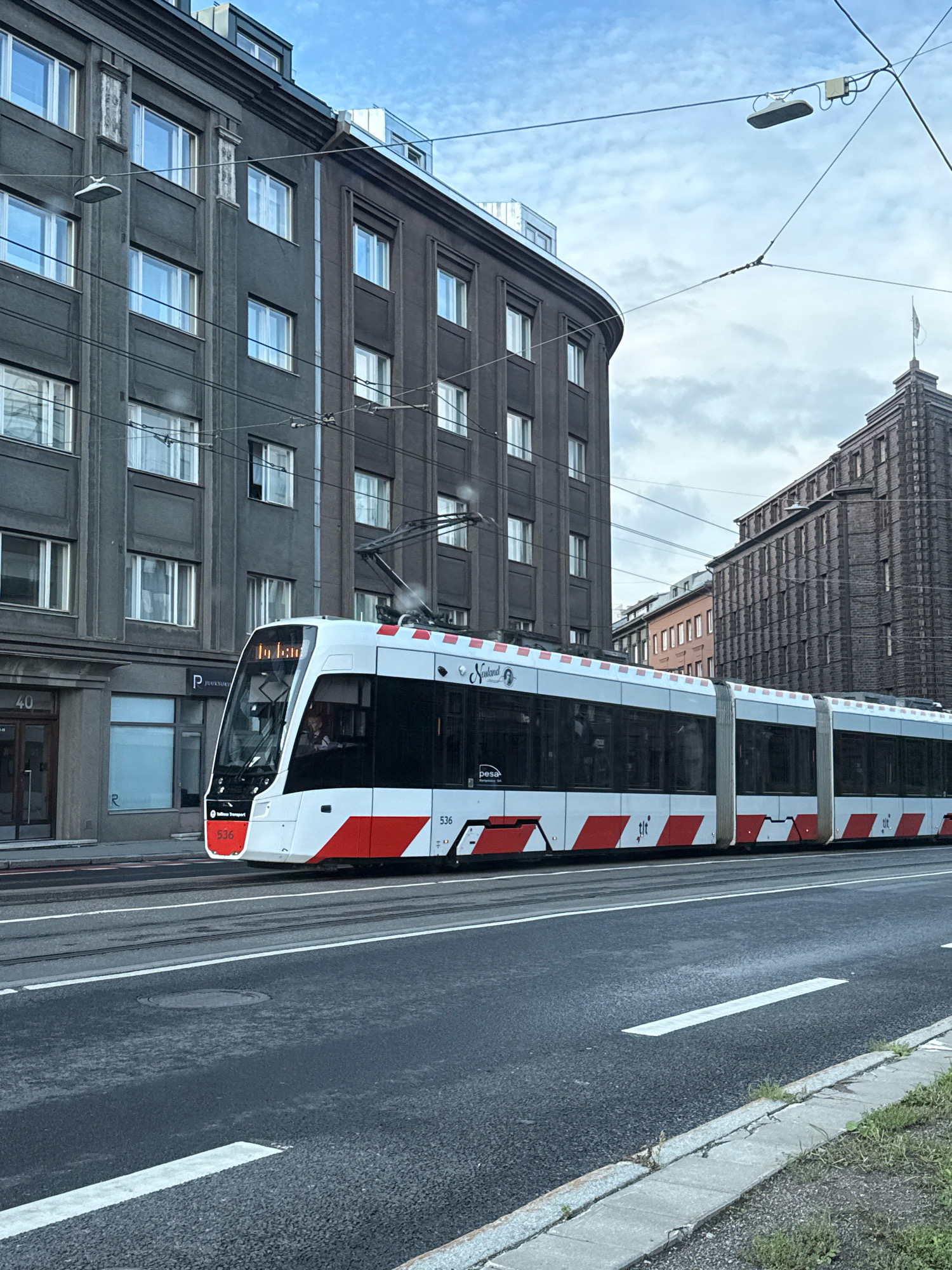 A modern white and red tram, number 536, is traveling on tram tracks in front of dark grey and brick buildings. The tram has a sleek design with large windows and appears to be in motion. The buildings are multi-story and have a somewhat dated architectural style. Overhead, power lines for the tram are visible against a cloudy sky. The street in the foreground is asphalt with white lane markings. A portion of a sidewalk with some greenery is visible at the bottom right of the image. The location appears to be an urban street in Tallinn, Estonia, given the visible signage on the tram which reads "Tallinna Transport" and the number "536" which corresponds to trams operating in Tallinn. The building on the left has the number "40" visible.