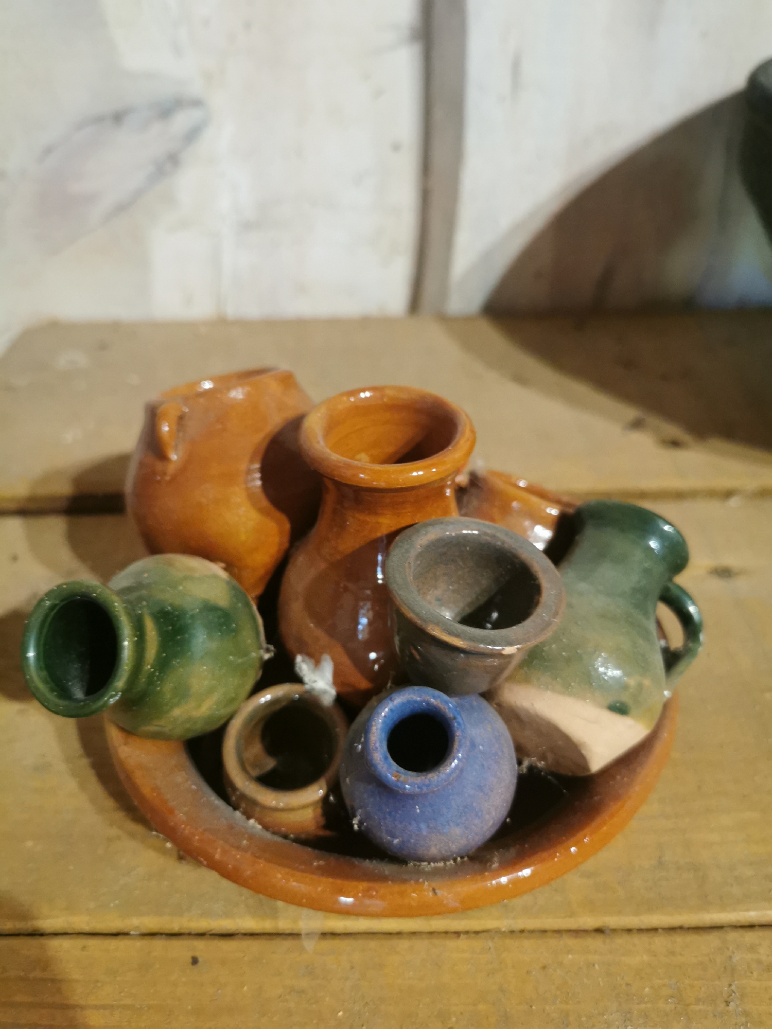 A collection of small, rustic ceramic vessels, including pitchers and vases, are arranged on a larger, shallow ceramic dish. The vessels vary in color, with shades of orange, green, blue, and brown. The largest orange pitcher is in the center, with a smaller orange pitcher to its right. A green pitcher with a handle sits to the right of the central orange pitcher. To the left is a rounded green vase, and in the foreground are two smaller, dark-colored cups or pots, one appearing blue and another a reddish-brown. The collection is displayed on a wooden surface, likely a table or shelf, in what appears to be an indoor environment with a textured, light-colored wall in the background. The lighting creates shadows, particularly on the wall behind the objects, suggesting a light source from the right. The overall impression is one of a still-life arrangement of handcrafted pottery, possibly for sale or display in a shop or personal collection in a rural setting.