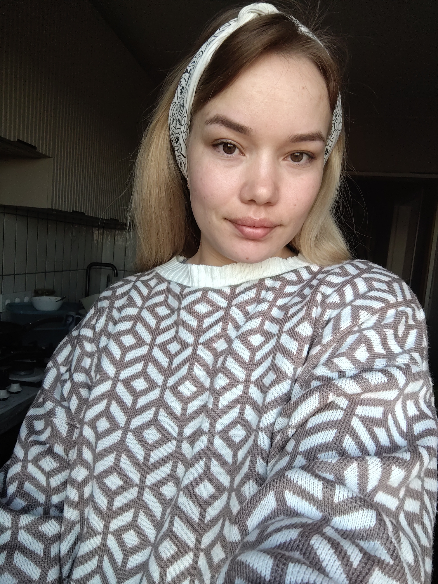 The image is a portrait of a young woman taken indoors, likely in a kitchen in Марковский, Russia. She is shown from the waist up, looking directly at the camera with a soft, relaxed expression and a faint smile. Her long, light brown hair is partially covered by a white bandana or headband adorned with a black floral or paisley pattern. Her eyes are brown, and her skin appears natural with minimal or no makeup. A small, delicate gold-colored earring is visible on her right ear.
She is wearing a cozy, knitted sweater with a distinctive geometric pattern of interlocking diamond or cube shapes in brown and white. The sweater has a high, ribbed white collar.
The background elements visible suggest a domestic setting, specifically a kitchen or utility room. To the left, there's a tiled wall, part of a sink with some dishes in a bowl, and what appears to be a stovetop with burners. The lighting indicates natural daylight, possibly from a window, casting some shadows. There is no visible text in the image.