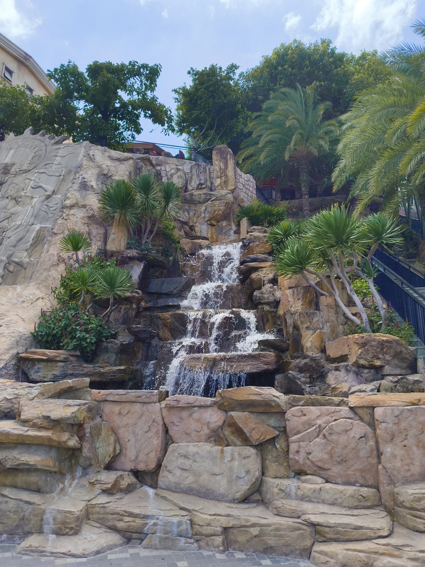 The image shows a multi-tiered artificial waterfall cascading down a rocky embankment. The rocks are varied in color and texture, with some appearing smooth and others rough. Lush green plants, including palm-like trees and leafy bushes, are integrated into the rock formations. At the top of the waterfall, a building with a light-colored facade and visible windows is partially obscured by trees. To the right, a dark metal staircase with railings ascends alongside the waterfall. The ground in front of the waterfall is paved with grey interlocking stones. The sky above is a light blue with scattered white clouds. This artificial waterfall is located in the Sun World Hon Thom Nature Park in Phu Quoc, Vietnam, specifically at the Aquatopia Water Park. There are no people visible in the image.