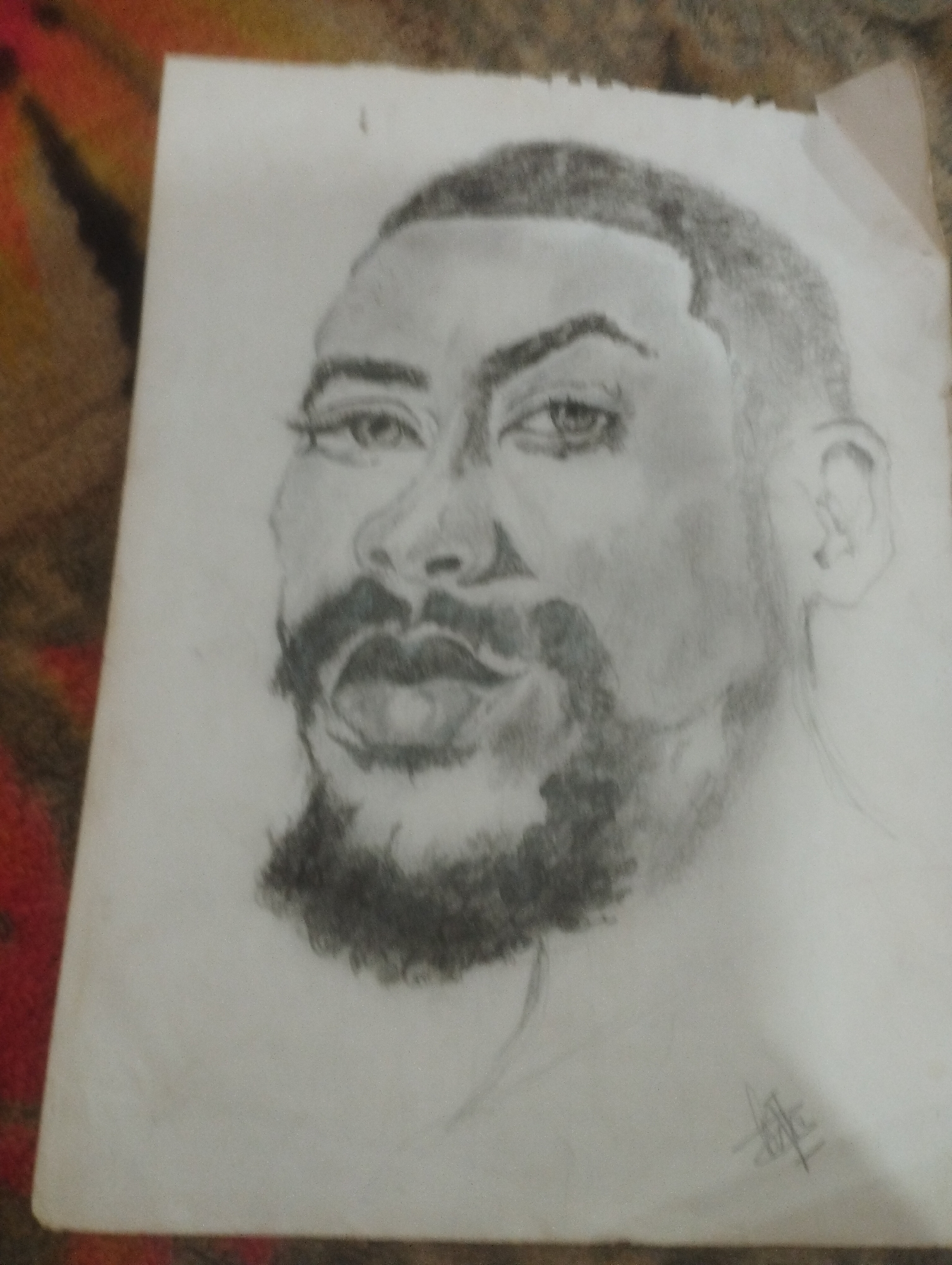 This is a pencil sketch of a Black man's face. He has short, dark hair, a full beard, and dark eyes. His head is tilted slightly to the left, and he is looking directly at the viewer. The sketch is rendered on white paper and appears to be framed or mounted on a patterned background. There is a signature in the lower right corner. The provided image does not contain enough information to determine the location in the city of Jos, Nigeria.