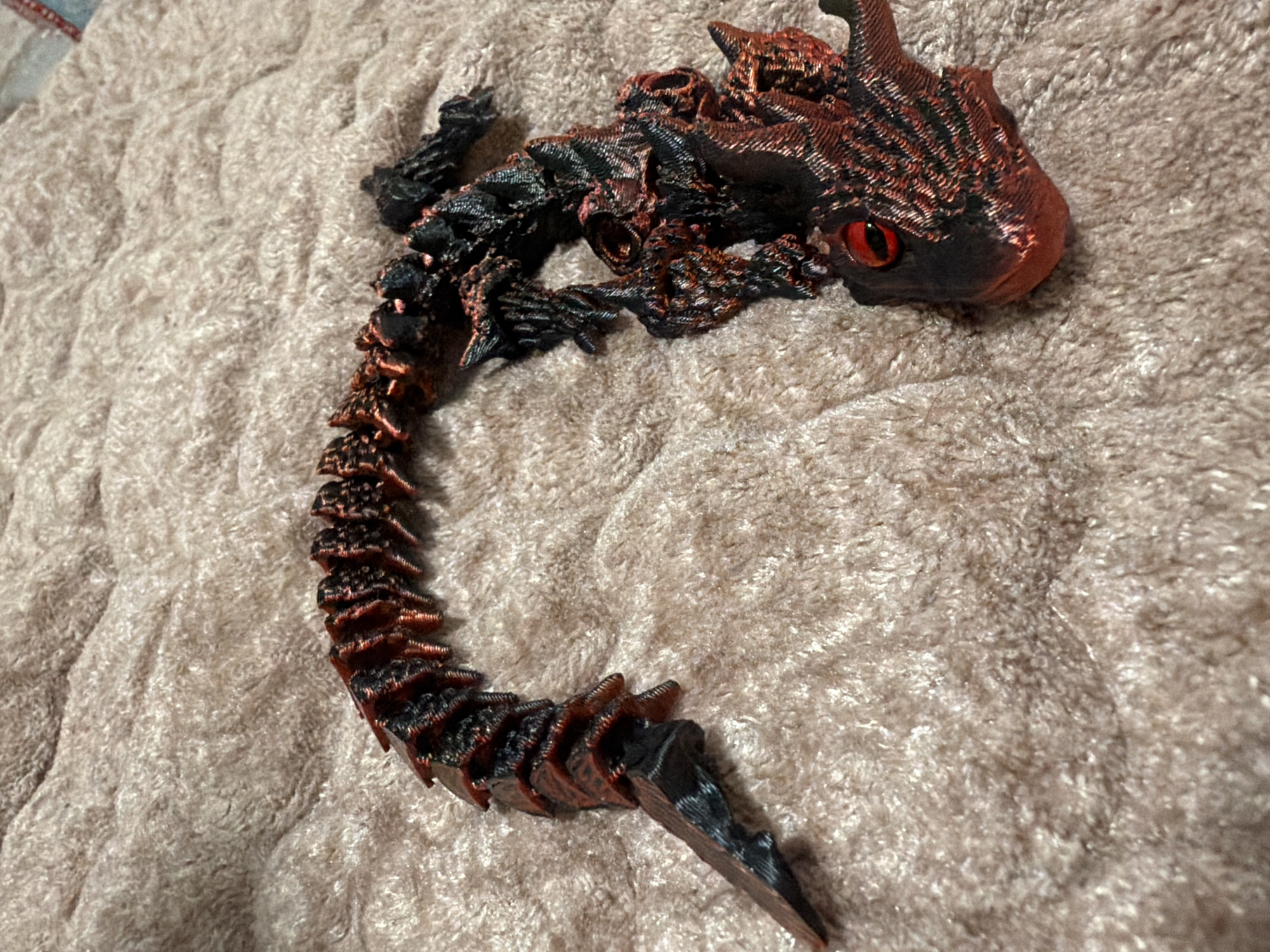A black and red segmented dragon toy is lying on a beige fluffy blanket. The dragon is coiled and its head is on the right side of the image, facing left. It has red eyes and intricate details on its body and head. The blanket appears to be a textured throw. The image quality is fair, with some noise and a shallow depth of field. The lighting is adequate but appears to be artificial. No people or animals are present in the image. The location is not specified.
