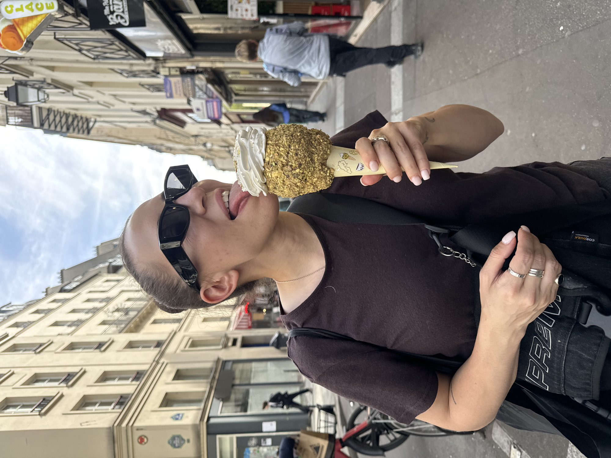 A woman with sunglasses is eating an ice cream cone on a city street. The ice cream cone is topped with white cream and what appears to be nuts. The buildings in the background suggest a European city, and a sign that says "Glace" indicates that the location is Paris, France. The woman is wearing a brown top, and two rings on her hand. The street has a bicycle and people walking in the background.