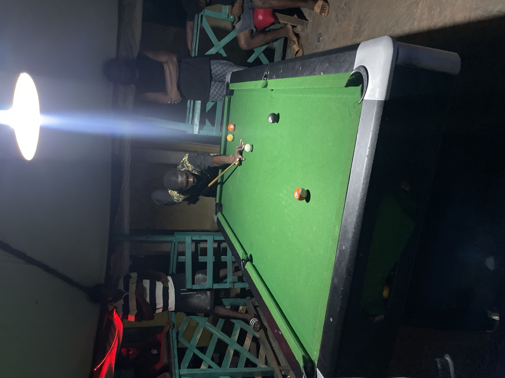 A man wearing a dark shirt with a patterned design plays pool on a green pool table in Lokoja, Nigeria. Another man wearing a striped shirt stands nearby, and two other people are sitting on a green bench in the background. There are a few billiard balls in view on the table. The image is illuminated by a bright light source. The environment appears to be somewhat outdoors, likely in a semi-enclosed space.