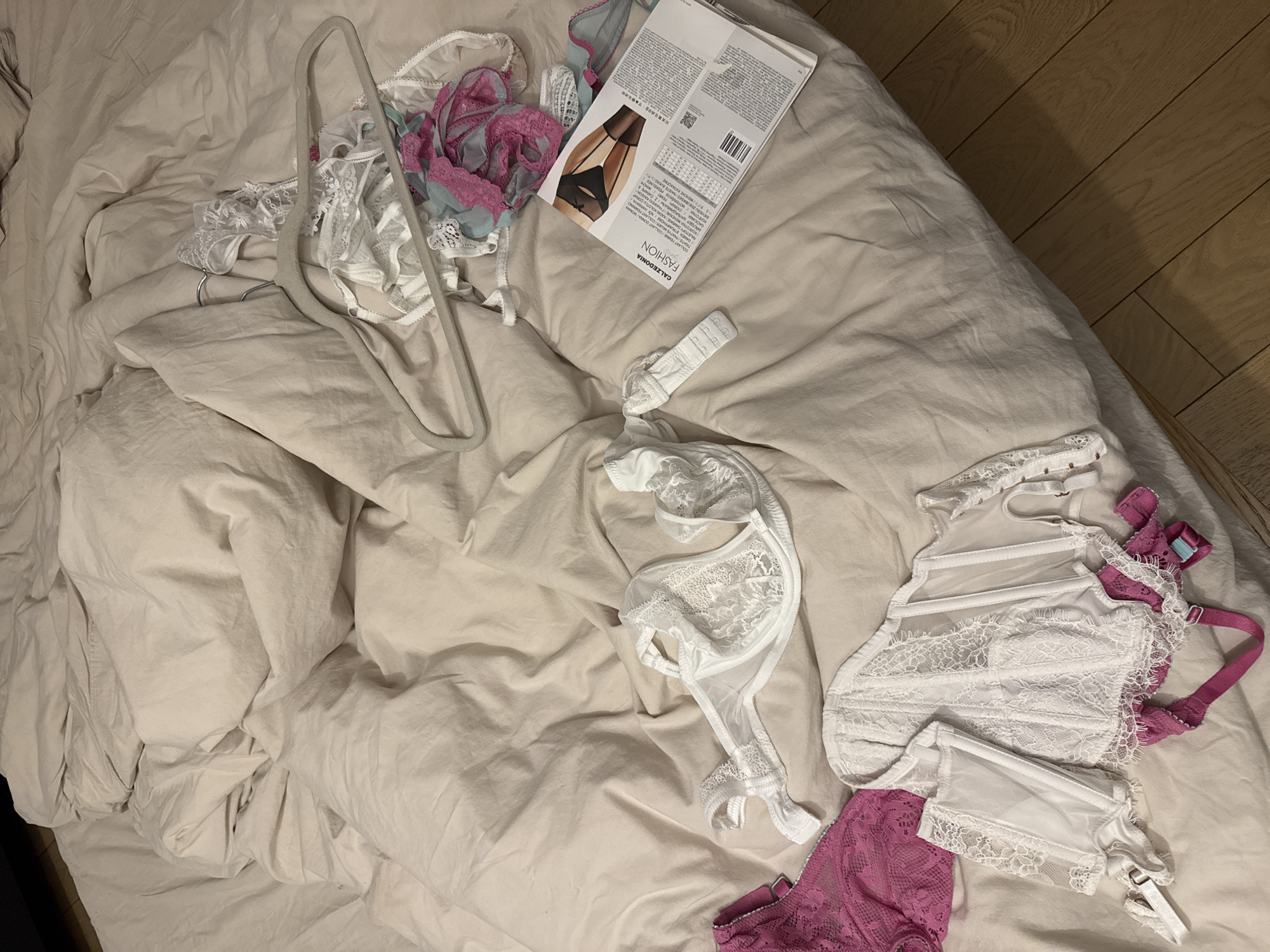 The image shows a disheveled bed with a beige duvet cover. Scattered on the bed are several items of women's lingerie, including bras, a garter belt, and what appears to be a hanger. A Calzedonia fashion panty package is also present. The scene appears to be indoors, possibly a bedroom. There are no visible people. The location in the city of Warsaw, Poland is impossible to determine from the image.