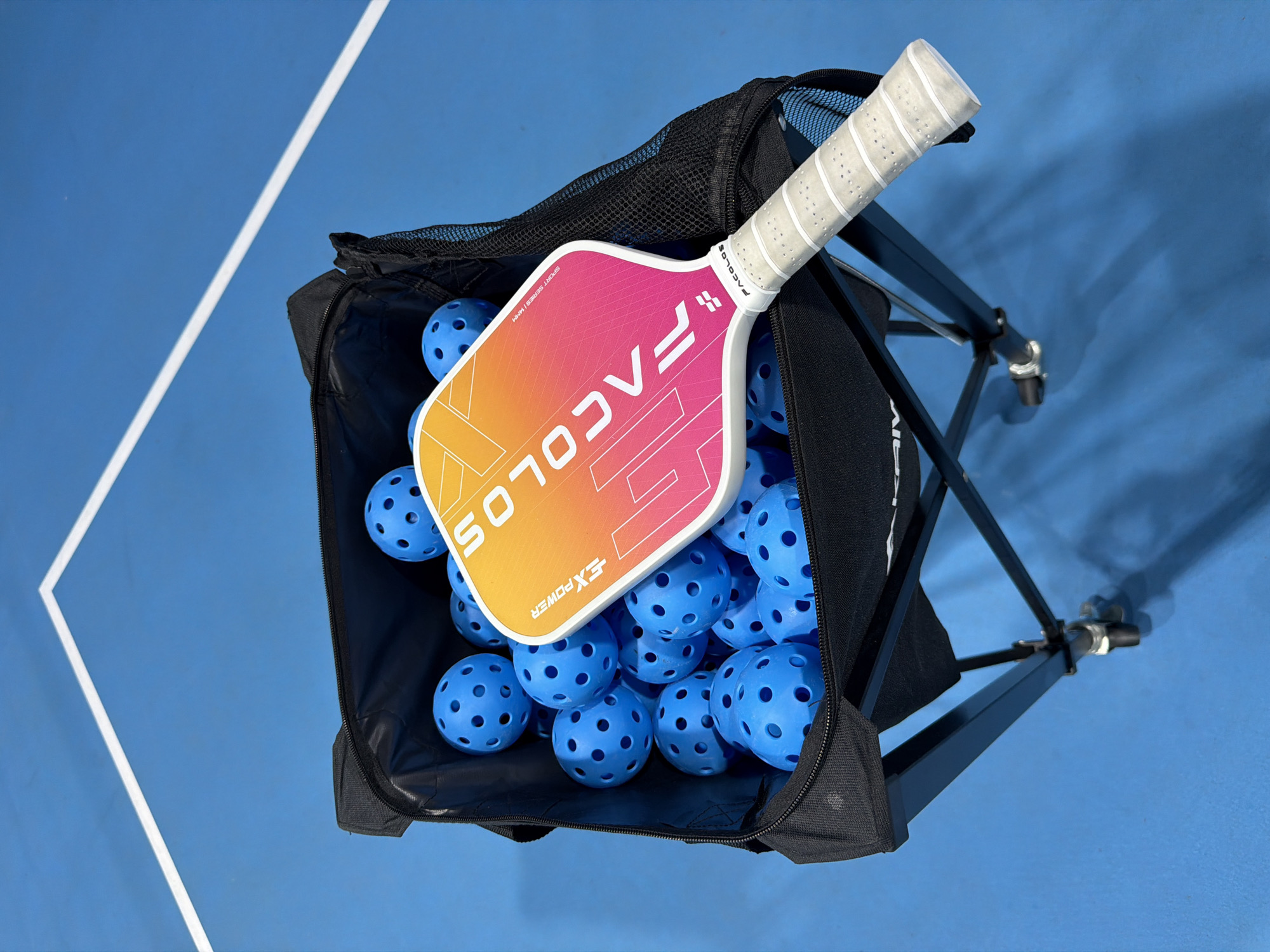 The image displays a pickleball set including a paddle and a bag full of pickleballs, placed on a blue pickleball court. The paddle has a gradient design from yellow to pink and features the "FACOLORS" logo. The pickleballs are bright blue and have holes. The setting appears to be outdoors on a sunny day, indicated by the bright lighting and shadows cast on the court. The blue court surface and the white boundary line are visible, suggesting a designated sports area. There is no explicit indication of Nha Trang, Vietnam in the image itself, but this context is provided separately. The overall impression is one of readiness for a game or practice session.