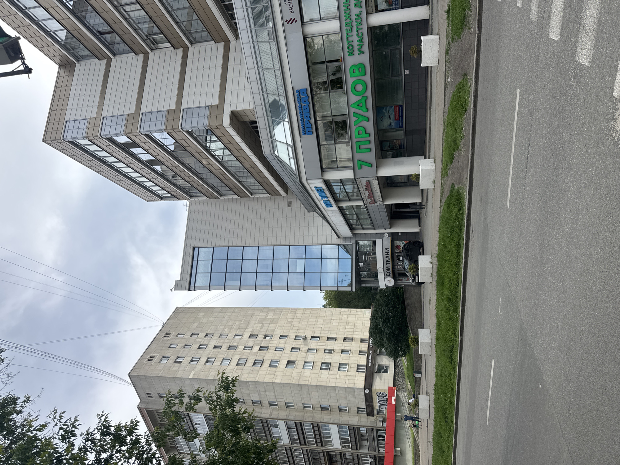 The image shows a street scene in Yekaterinburg, Russia. In the foreground is a two-lane asphalt road with a white line marking the center and a white pedestrian crossing. A wide grassy median separates the road from a sidewalk. Behind the sidewalk are several buildings. The closest building to the right is a modern, multi-story building with a glass facade and tan and white paneling. It features a prominent green sign that reads "7 ПРУДОВ" and smaller text indicating "КОТТЕДЖНЫЕ УЧАСТКИ, ДОМ". Next to this, there is another sign with the text "ДОМ ТКАНИ". To the left of this modern building is a taller, gray, concrete apartment building with many windows. Further to the left, a portion of another building with a red facade is visible. Several trees and bushes are planted along the sidewalk and around the buildings. Two individuals, an adult and a child, are walking on the sidewalk in front of the gray apartment building. A black car is parked on the street in front of the "ДОМ ТКАНИ" store. The sky is overcast. Overhead, several electrical wires are suspended between the buildings.