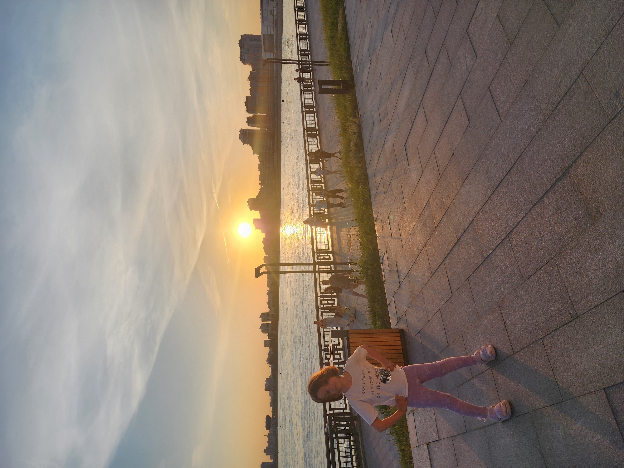 Here is a description of the image:
A young girl stands on a paved walkway near a body of water at sunset. The walkway is made of large, rectangular stone tiles. The girl is wearing a white t-shirt and light purple leggings. In the background, across the water, are city buildings silhouetted against the bright setting sun. A metal railing and grass separate the walkway from the water's edge. Several people can be seen walking or standing near the railing in the background.