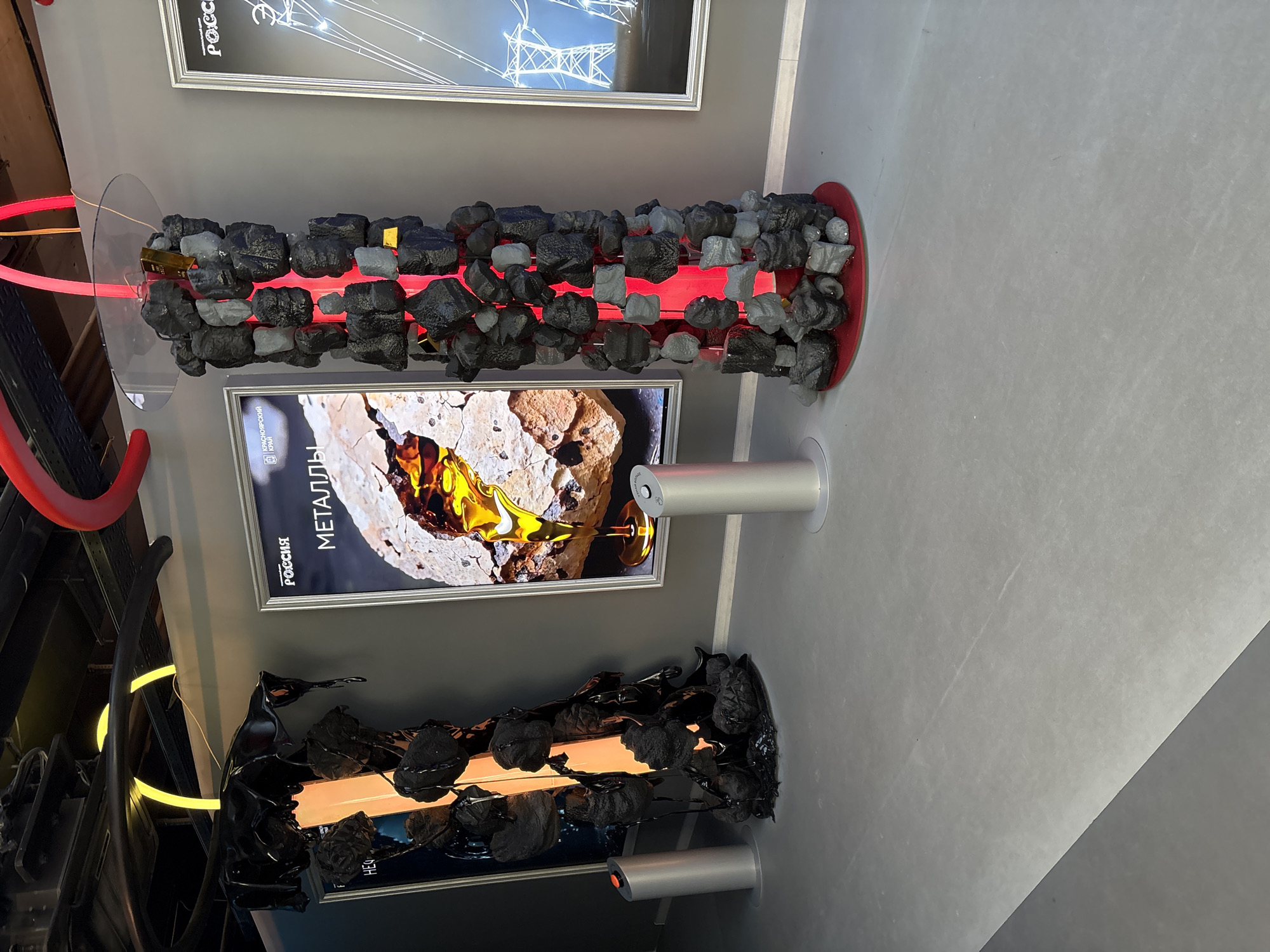 The image shows an indoor exhibit in an unknown location in the city of Unknown, Unknown. The scene features three vertical displays. The top display is illuminated with a red neon light running through a column of black and gray rocks. The middle display has a framed advertisement or information panel featuring the word "МЕТАЛЛЫ" (metals) in Cyrillic. Below this is a base with a silver cylinder. The bottom display appears to be a similar setup, with a frame displaying an orange light behind a black and gray structure reminiscent of rocks. This display also has a base with a silver cylinder. A fourth display is visible on the far left, featuring red and yellow neon lights near a dark structure. The background is a plain gray wall, and the floor is a light gray.