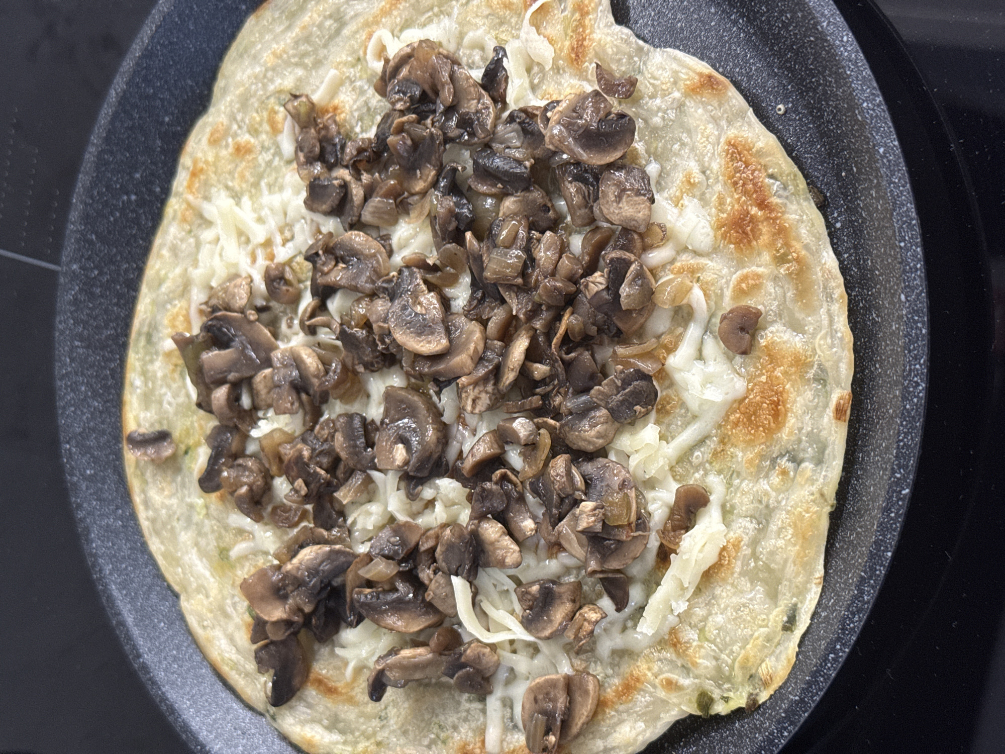 A close-up, overhead shot shows a mushroom and cheese quesadilla cooking in a black, non-stick pan. The quesadilla is golden brown and appears to be made with a thin, layered tortilla, possibly a paratha. Shredded white cheese is melted over the tortilla, and a generous topping of sauteed mushrooms, sliced and mixed with what appears to be finely chopped onions, covers the cheese. The pan is on a black stovetop, which is the only visible element of the environment. There are no people or other objects in the frame. The image captures a culinary moment, likely in a kitchen. There is no information about the location in Tomilino, Russia, that can be inferred from this image.