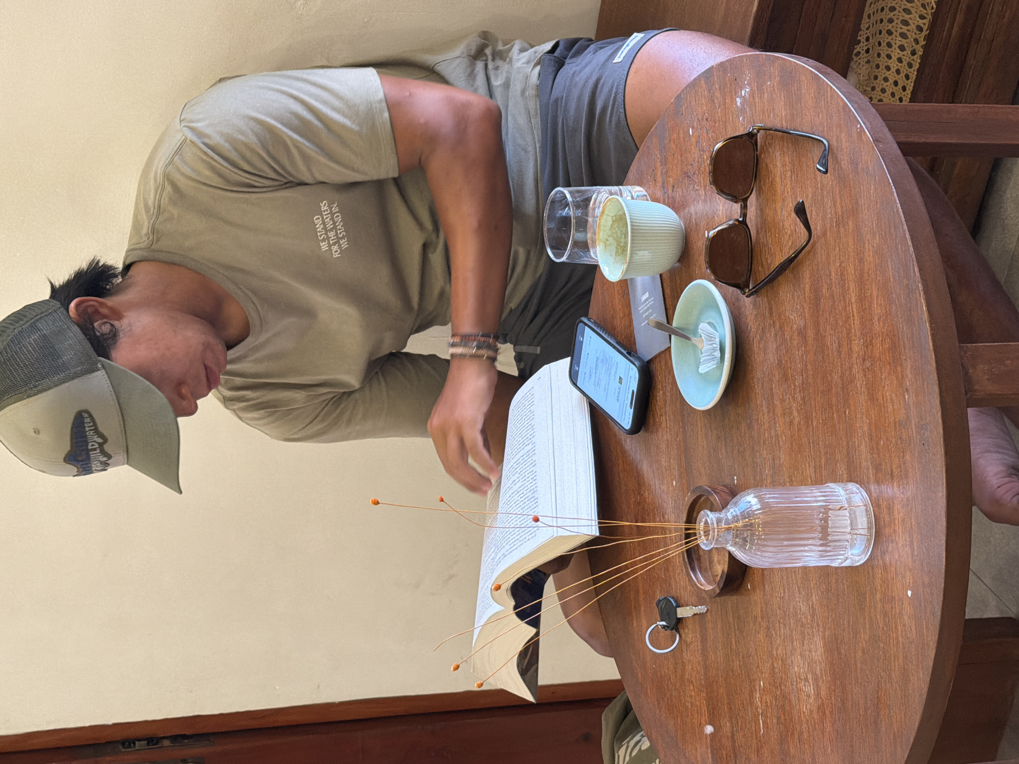 A man is seated at a round wooden table, seemingly engaged in reading a book or looking at his smartphone. He wears a light khaki t-shirt with text, dark shorts with a small white tag, and a grey and white trucker cap with text. A multi-strand beaded bracelet adorns his right wrist.
On the wooden table, an open book lays alongside a black smartphone displaying content. Nearby are two used cups, one clear glass and one ridged light blue-green ceramic, with a matching saucer and spoon. A pair of brown tortoiseshell sunglasses rests further back on the table. A small ribbed glass vase holds delicate, wired decorative elements with orange tips. A single key on a metal keyring is also visible on the table. In the background, a light-colored wall is visible, along with glimpses of wooden furniture. A bare foot can be seen in the bottom right of the frame.
Notable text includes: "WE STAND FOR THE WATERS WE STAND IN." printed on the man's t-shirt. His cap has partially visible text, possibly "WILDWATERS." A small tag on his shorts reads "LAGUNA," and a label on the table next to a cup shows "LABORA." The scene conveys a relaxed, casual atmosphere, likely during the daytime.
