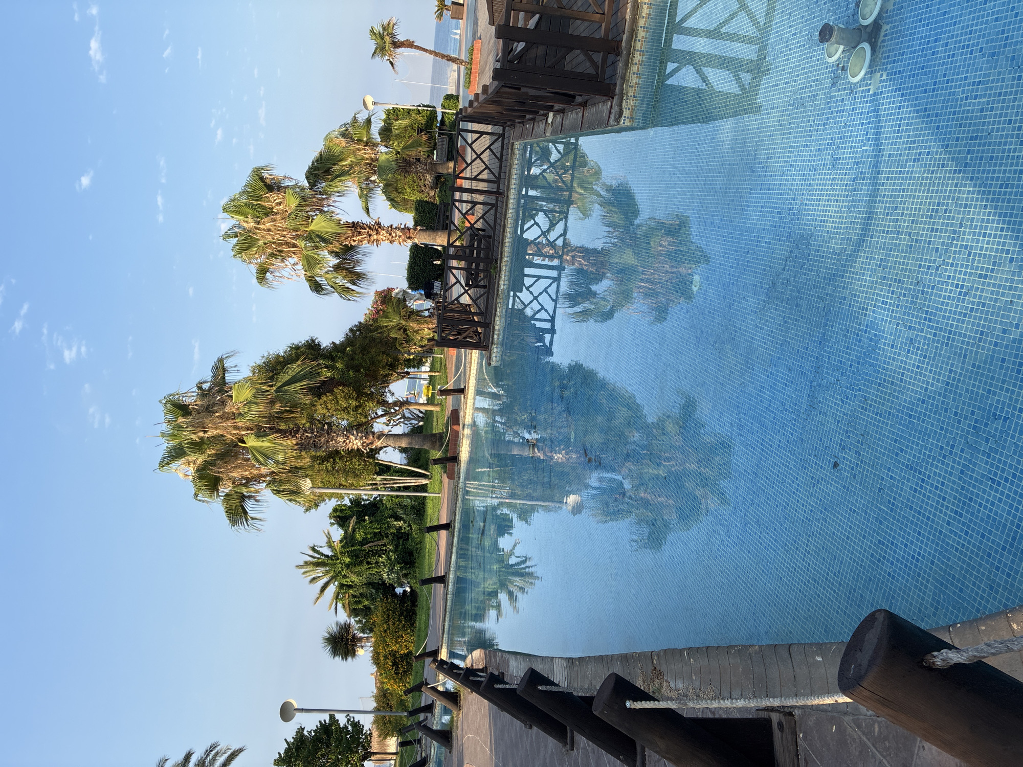 The image depicts an outdoor swimming pool with clear blue water, tiled with small blue squares. The pool is bordered by a paved walkway and a wooden railing. Several palm trees are visible, some reflected in the water. In the background, there is a glimpse of the sea and a clear blue sky with scattered clouds. The setting appears to be a resort or public area in Alanya, Turkey, likely during daytime.