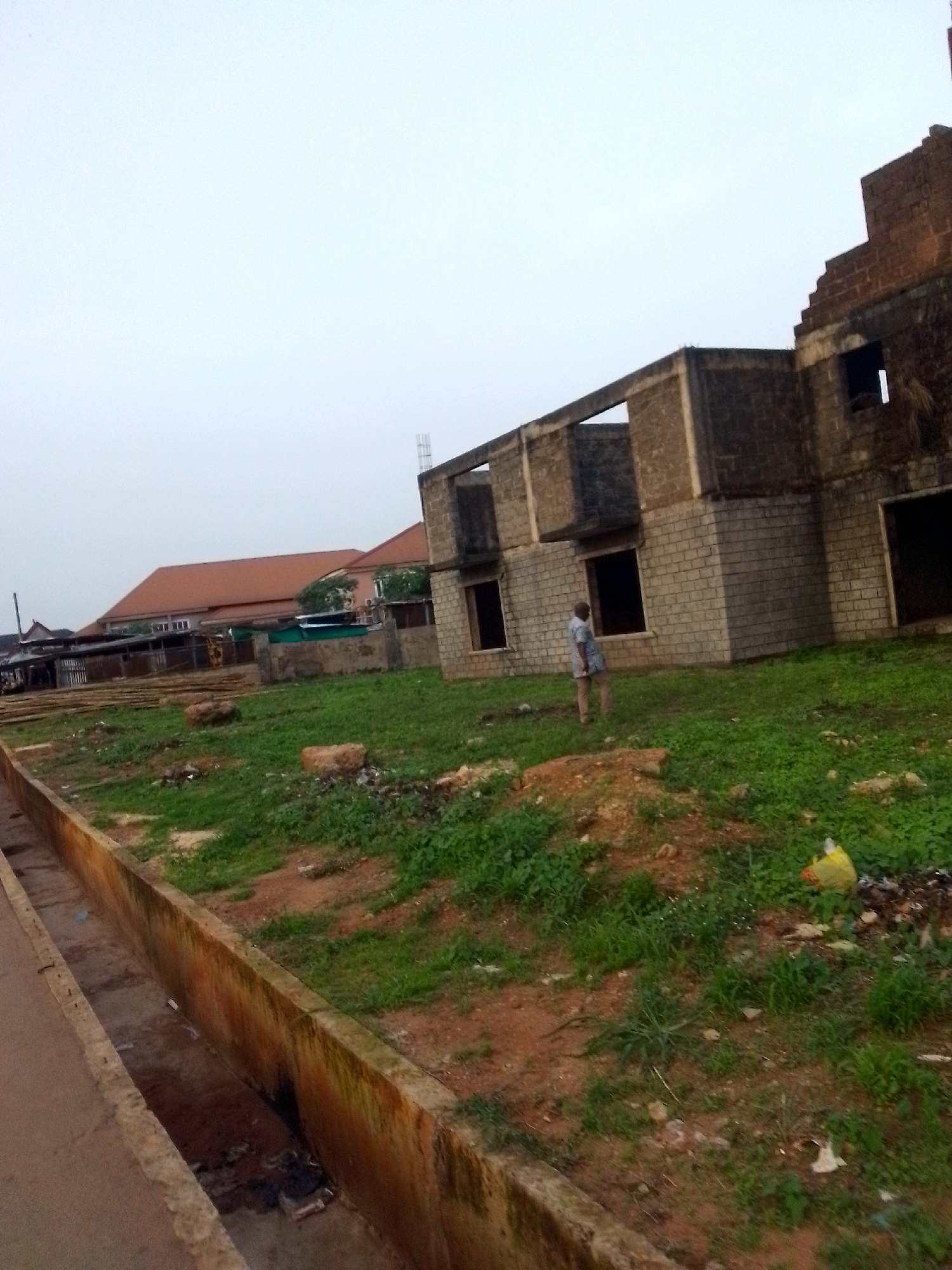 The image shows an unfinished two-story building in what appears to be an undeveloped area. The building is constructed of concrete blocks, with open window and door frames. A man stands on the grassy and dirt-covered land in front of the structure. To the left, a ditch runs along a road. In the background, there are other buildings, including one with a red roof. Debris, including a yellow plastic bag, is scattered on the ground. Given the prompt, the scene is assumed to be in Orlu, Nigeria. The sky is overcast.