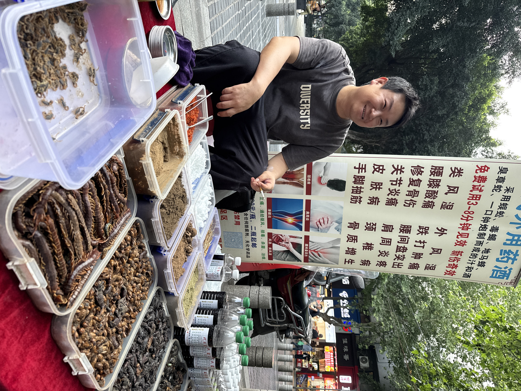 The image depicts a street vendor in Beibei, China, selling traditional Chinese medicine. The vendor, a middle-aged man with a gray t-shirt with “DIVERSITY” in white letters, sits behind a table displaying various ingredients in clear plastic containers, including dried insects and powders. The ingredients are displayed atop a red cloth. A sign behind the vendor advertises the medicinal properties of the products, promising relief from ailments such as rheumatoid arthritis, lower back pain, and skin conditions. Bottles of liquid and stacks of gray stones are also displayed. A red moped is parked behind the vendor, and storefronts are visible in the background. The location is on a paved area in front of storefronts, likely a pedestrian zone or sidewalk in Beibei, China.