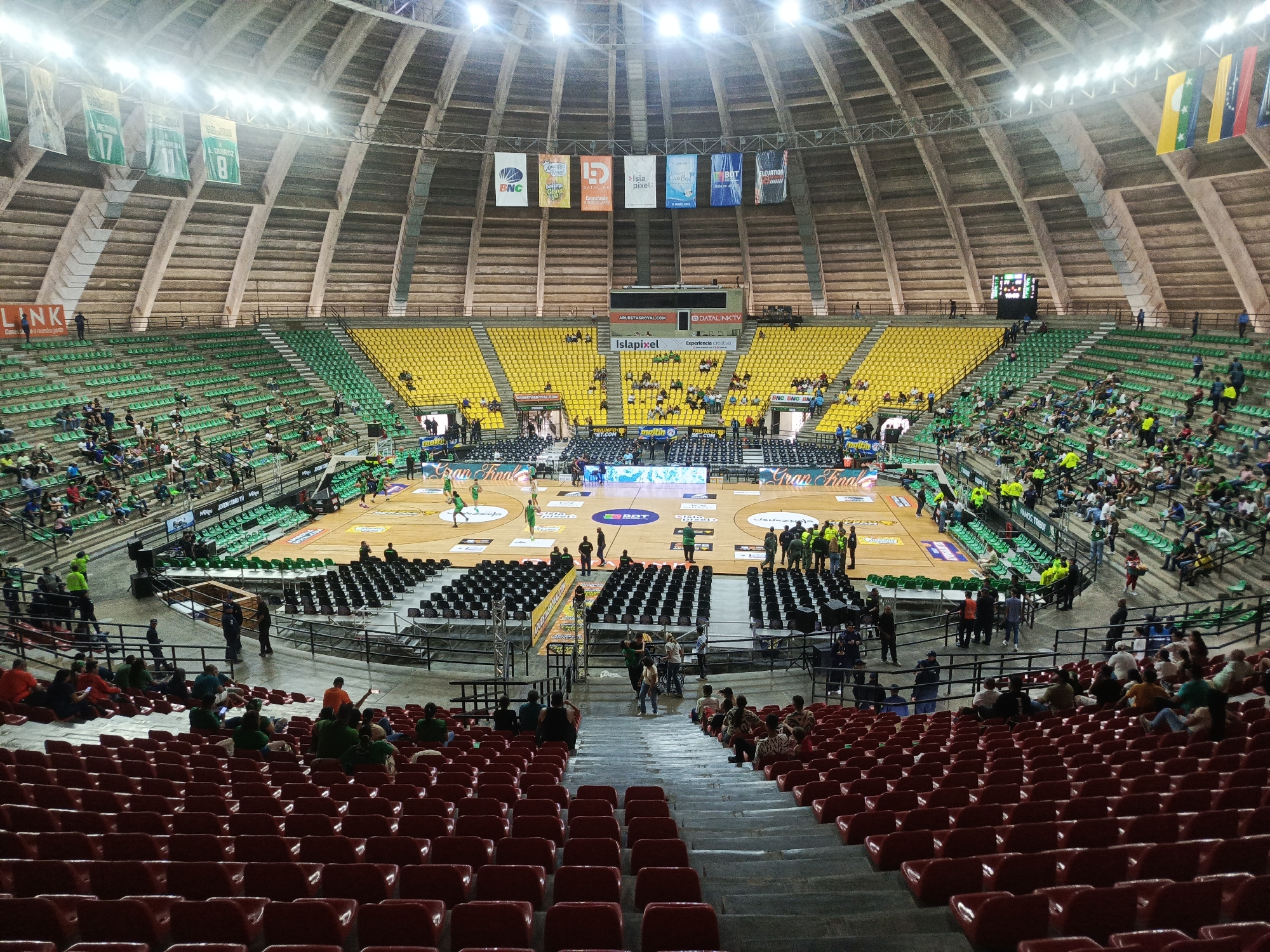 The image captures a wide view of an indoor basketball arena during a game, likely in La Asunción, Venezuela. The arena features tiered seating in green and yellow, with many seats empty. A basketball court is visible with players in green uniforms in action. Banners hang from the ceiling, displaying various logos and years. Spectators are scattered throughout the stands, some actively watching the game, others conversing. The architecture of the arena is characterized by a domed ceiling and concrete supports. The overall atmosphere appears to be that of a sporting event with a moderate crowd.
