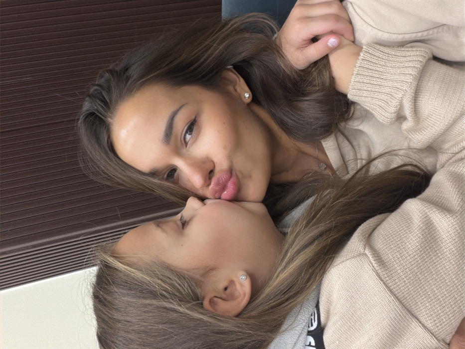 A close-up indoor portrait features an adult female and a young female child. The child, with light brown hair and wearing a beige hooded sweater, is positioned on the left, with her profile facing right, pressing her lips against the adult's left cheek in a kiss. She has a small, circular stud earring in her visible left ear. The adult female, with long light brown hair and also wearing a beige hooded sweater, is positioned on the right, looking directly towards the viewer with her lips puckered. She wears a small stud earring in her right ear and a delicate silver-colored necklace with a small charm. The child's right hand is visible, resting on the adult's left shoulder. The background consists of a dark brown, vertically slatted wall on the right and a lighter, solid wall section on the upper left. No specific identifying features are present in the image to confirm the location as Krasnoyarsk, Russia.