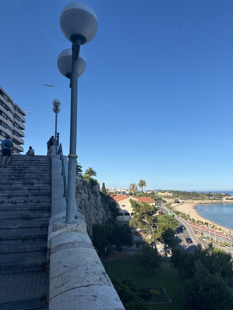 The image captures a view from a high vantage point in Tarragona, Spain, looking towards the coastline. In the foreground, there is a set of stone steps and a low stone wall. Several people are ascending the steps, while others are seated near the top. Two prominent lampposts with spherical lights stand beside the steps.
Beyond the wall, a cliff descends to a road that runs parallel to the beach. Buildings, trees, and a grassy area are visible between the cliff and the road. In the background, a sandy beach stretches along the coastline, meeting the blue sea. The sky is clear and bright. Several birds are flying in the sky.