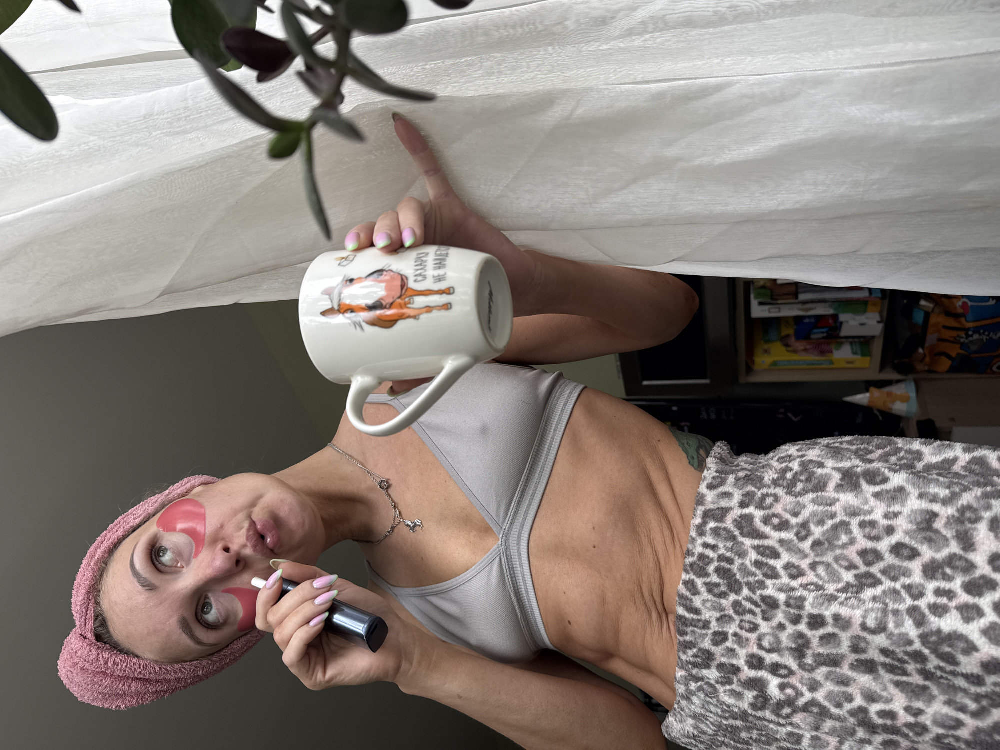 A woman with a pink towel turban and pink under-eye patches holds a vape pen to her lips and a white mug with a horse design in her right hand. She is wearing a grey sports bra and leopard-print pajama pants. The background shows a white sheer curtain and a bookshelf. The image appears to have been taken indoors, possibly in a residential setting.